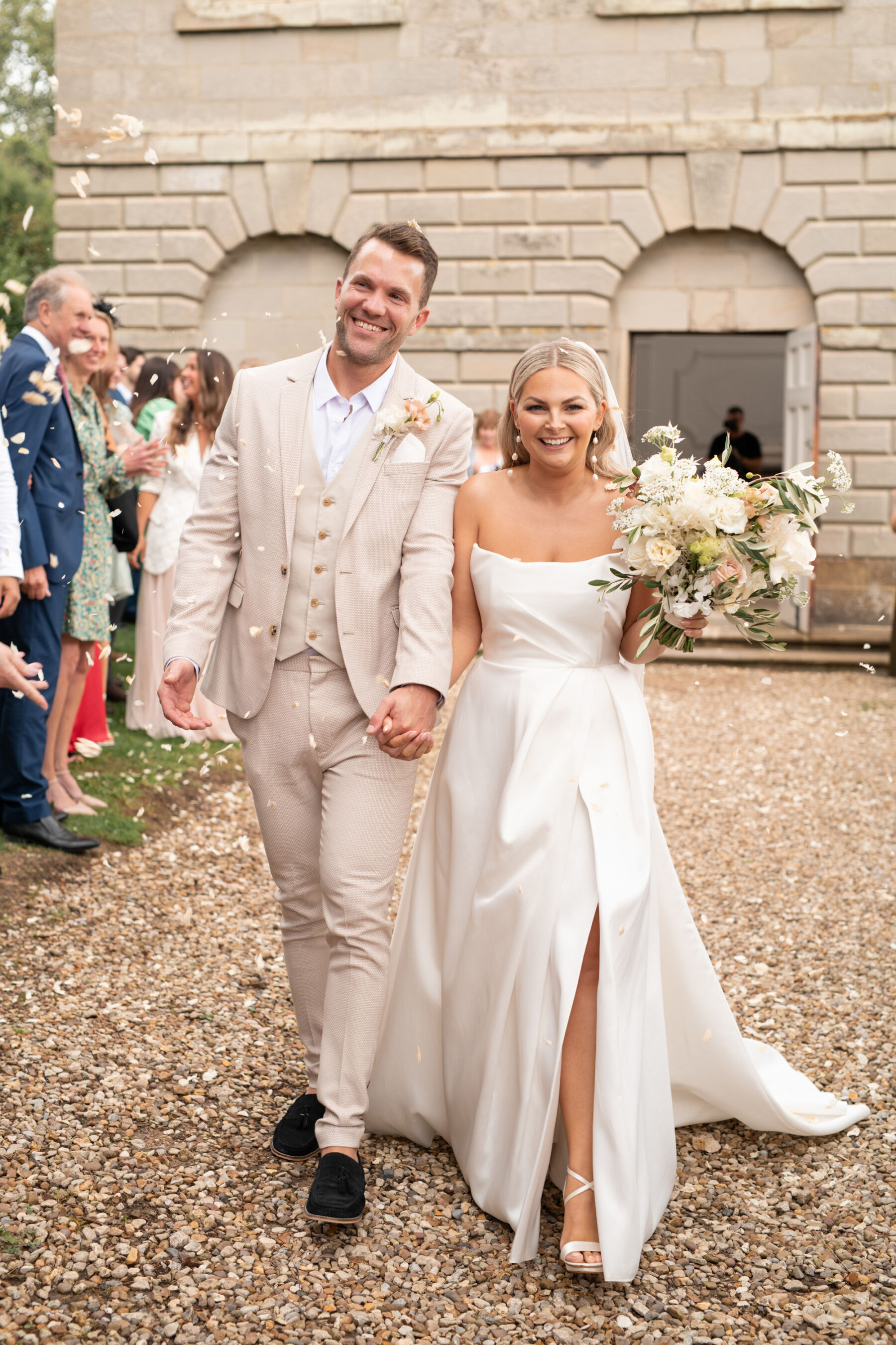 Bride wearing an Aleena Leena wedding dress with a thigh slit. Groom in pale suit. Newlyweds exiting church to a confetti shower.