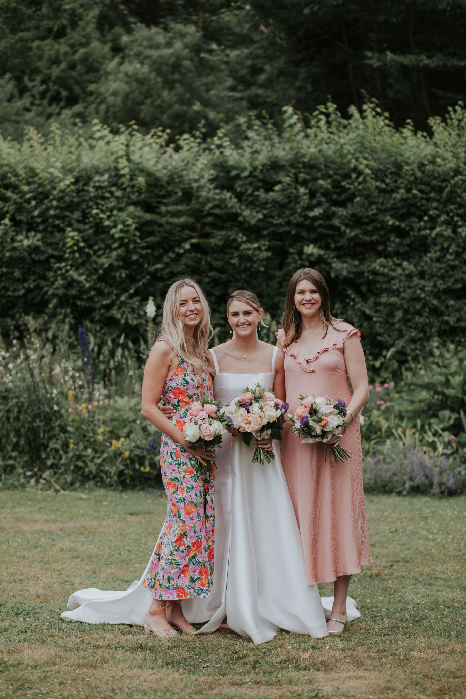 Bride in a Khya wedding dress standing on the grass with a bridesmaid in pink and floral dresses standing either side of her.