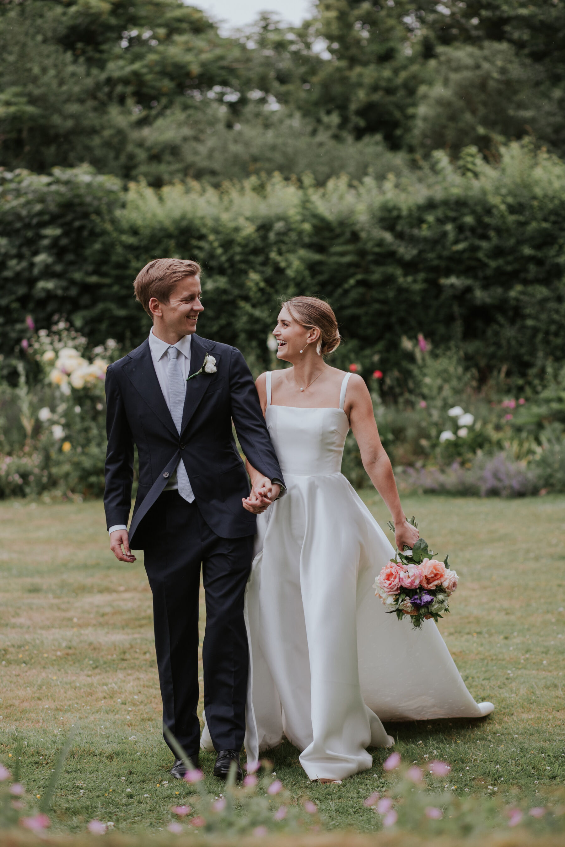 Groom in navy blue suit from Suit Supply holding hands with bride in a square neck Khya wedding dress as they walk through an English country garden.
