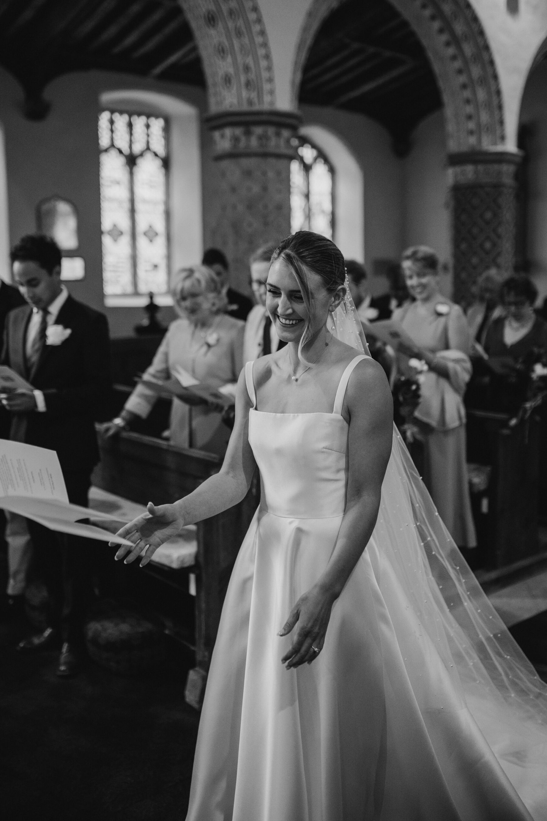 Bride smiling in church during the wedding ceremony. Bride wears a long polka dot veil and square neck dress by Khya.