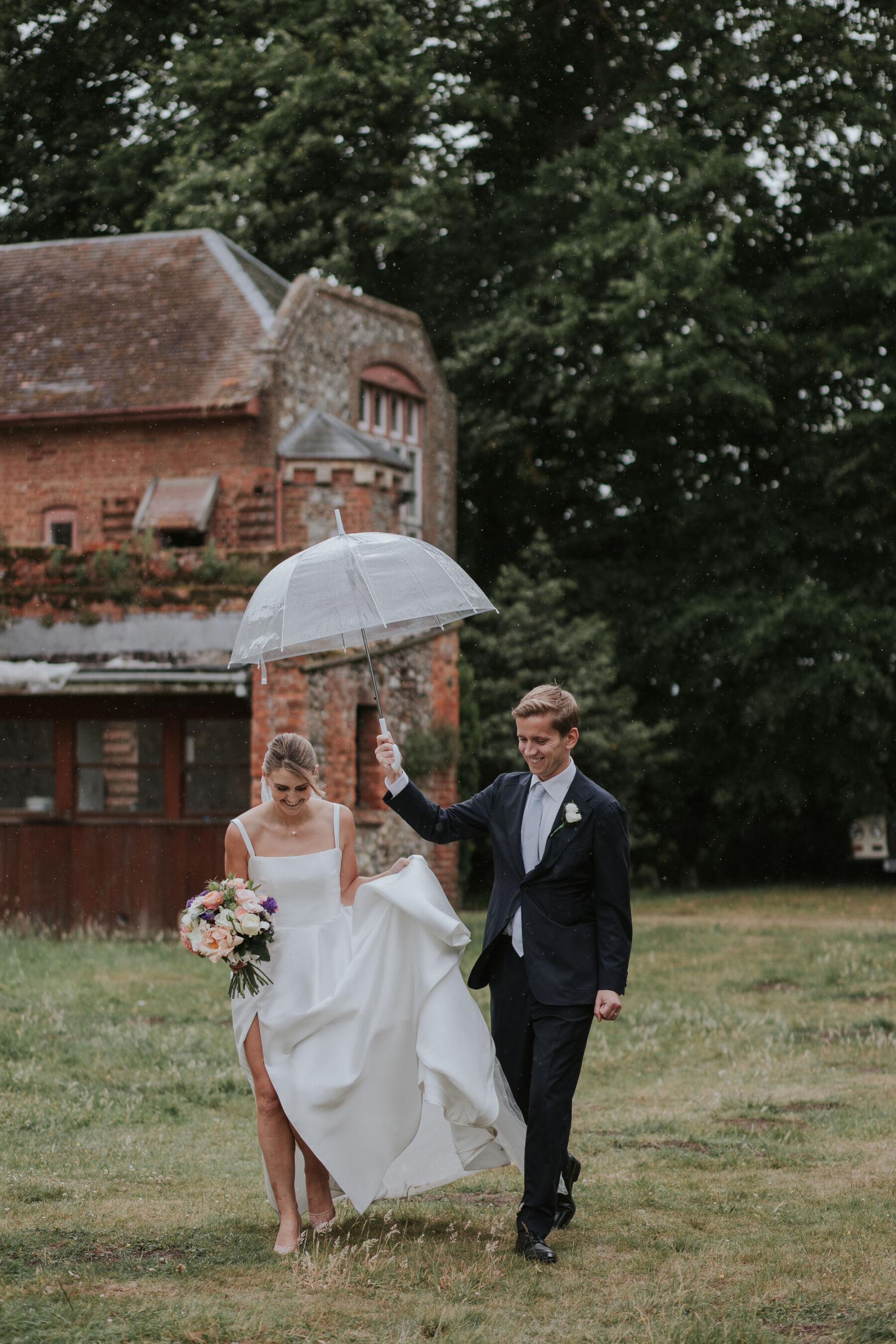 Groom holding a clear umbrella over his bride as they walk through the grass.