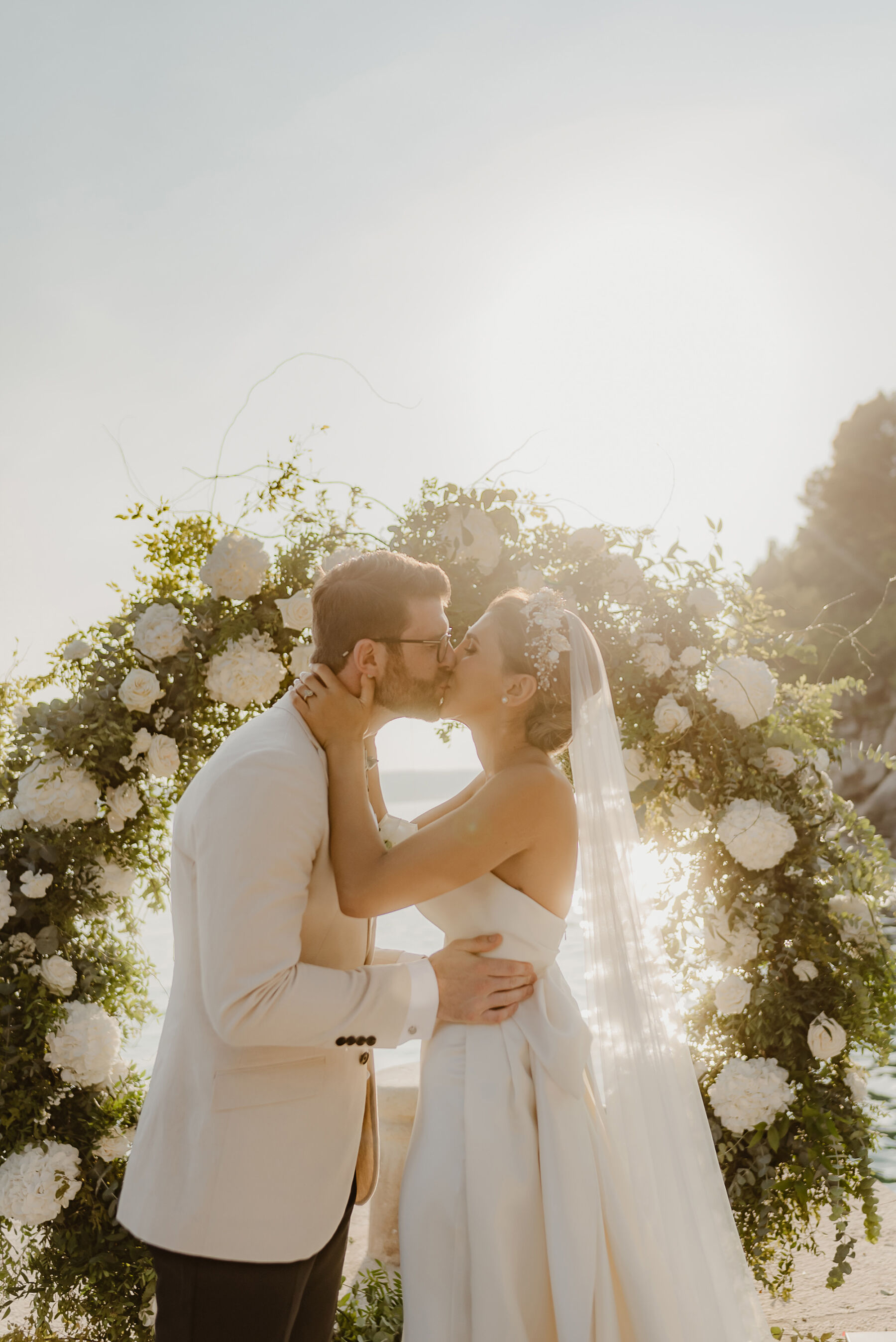 Sarah in Eva Lendel for her Classy & Contemporary Wedding in Croatia 30 Bride and groom kissing by a floral arch of white flowers