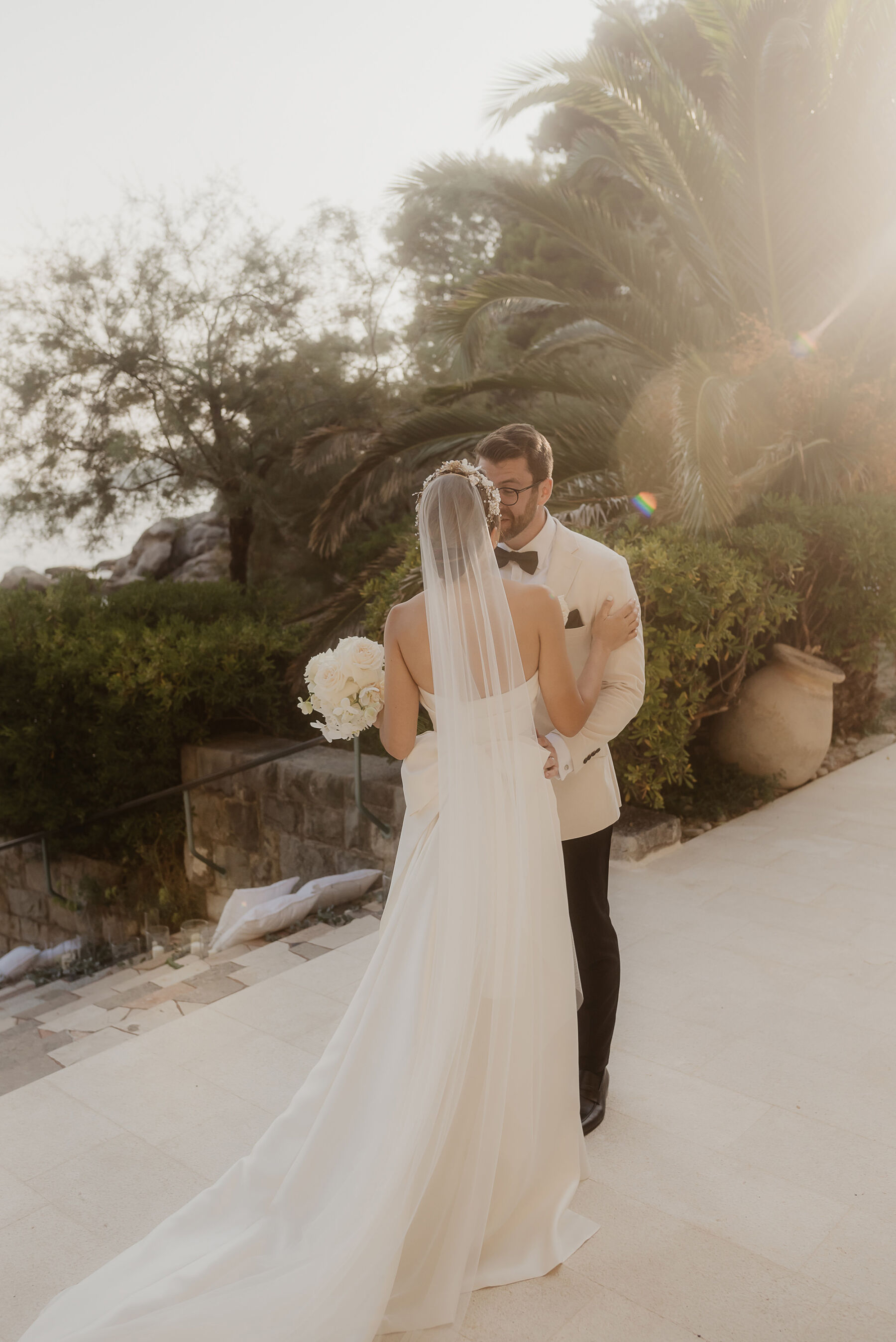 Groom in white tux kissing bride in minimalist wedding dress and long veil carrying a bouquet of all white flowers