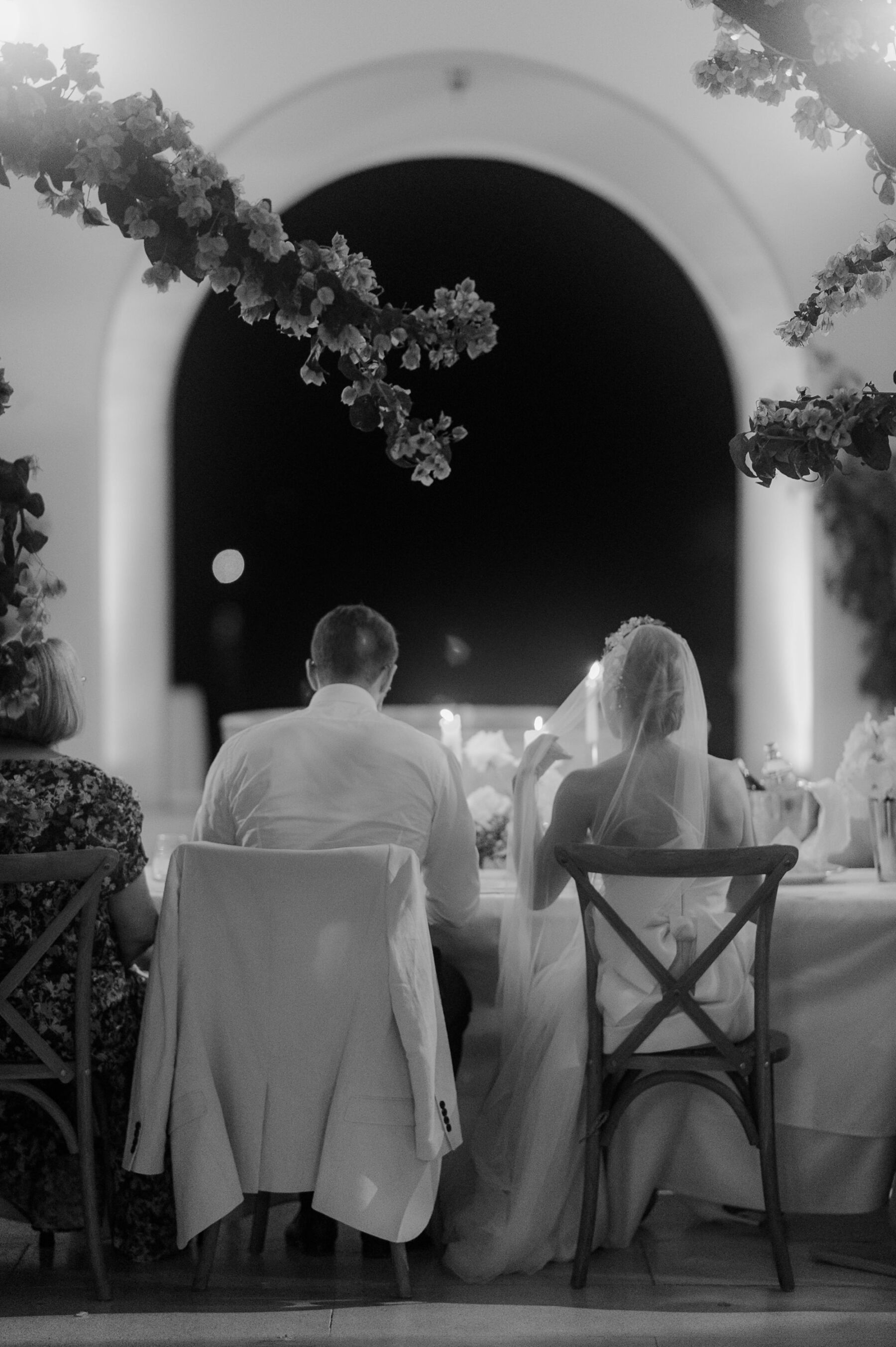 Shot from behind of bride and groom sitting next to one another on cross back wooden chairs with an archway head of them at night time.