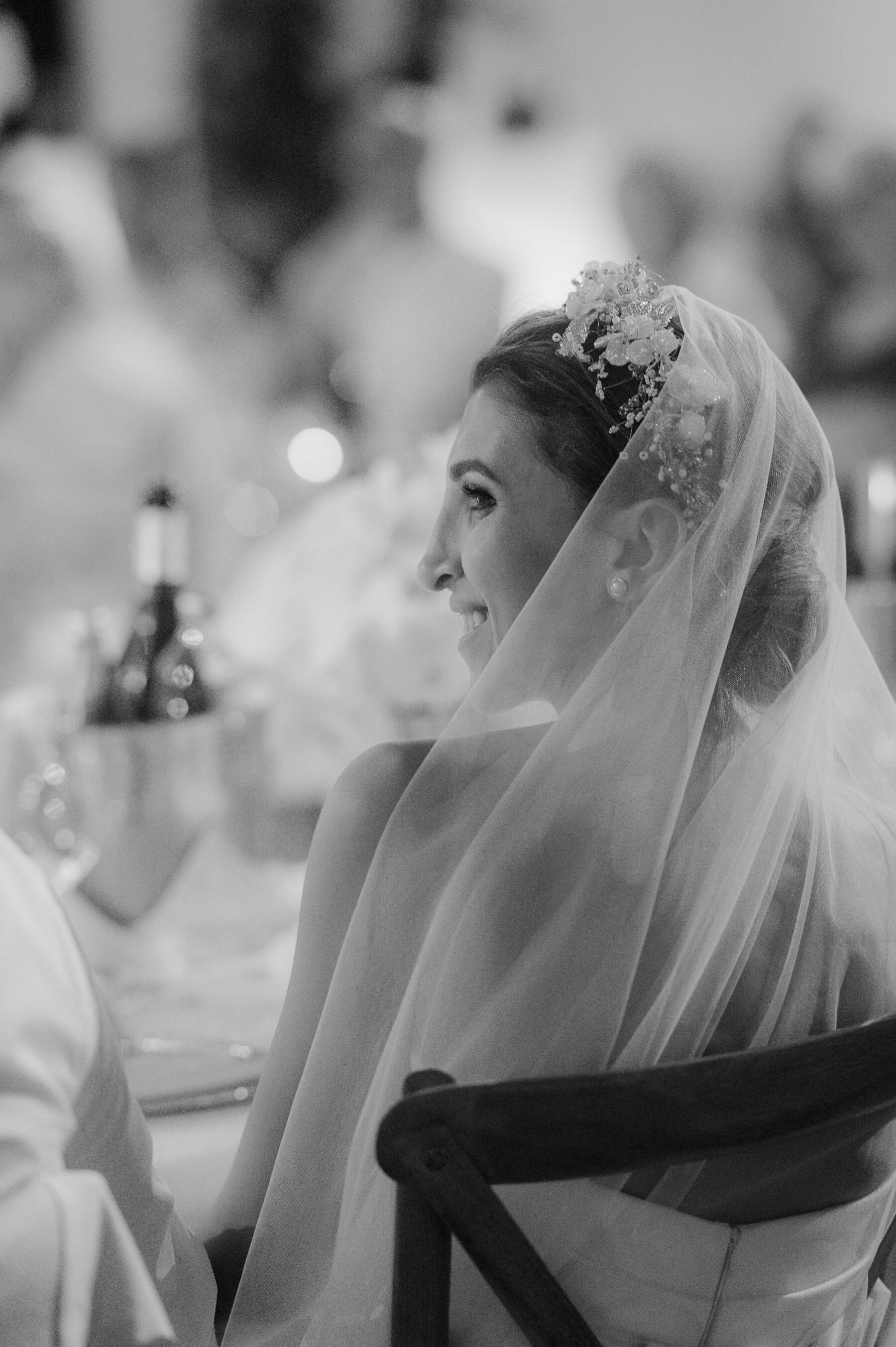 Black and white profile shot of bride in a tulle veil and mother of pearl headpiece seated at the wedding reception.