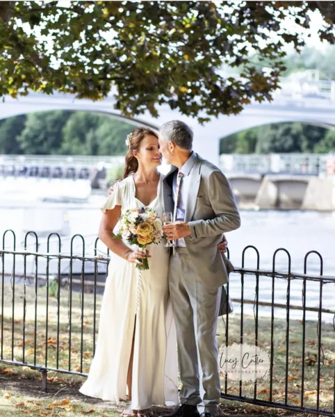 A bride & groom are stood by the River Thames. They are kissing and holding a glass of champagne.