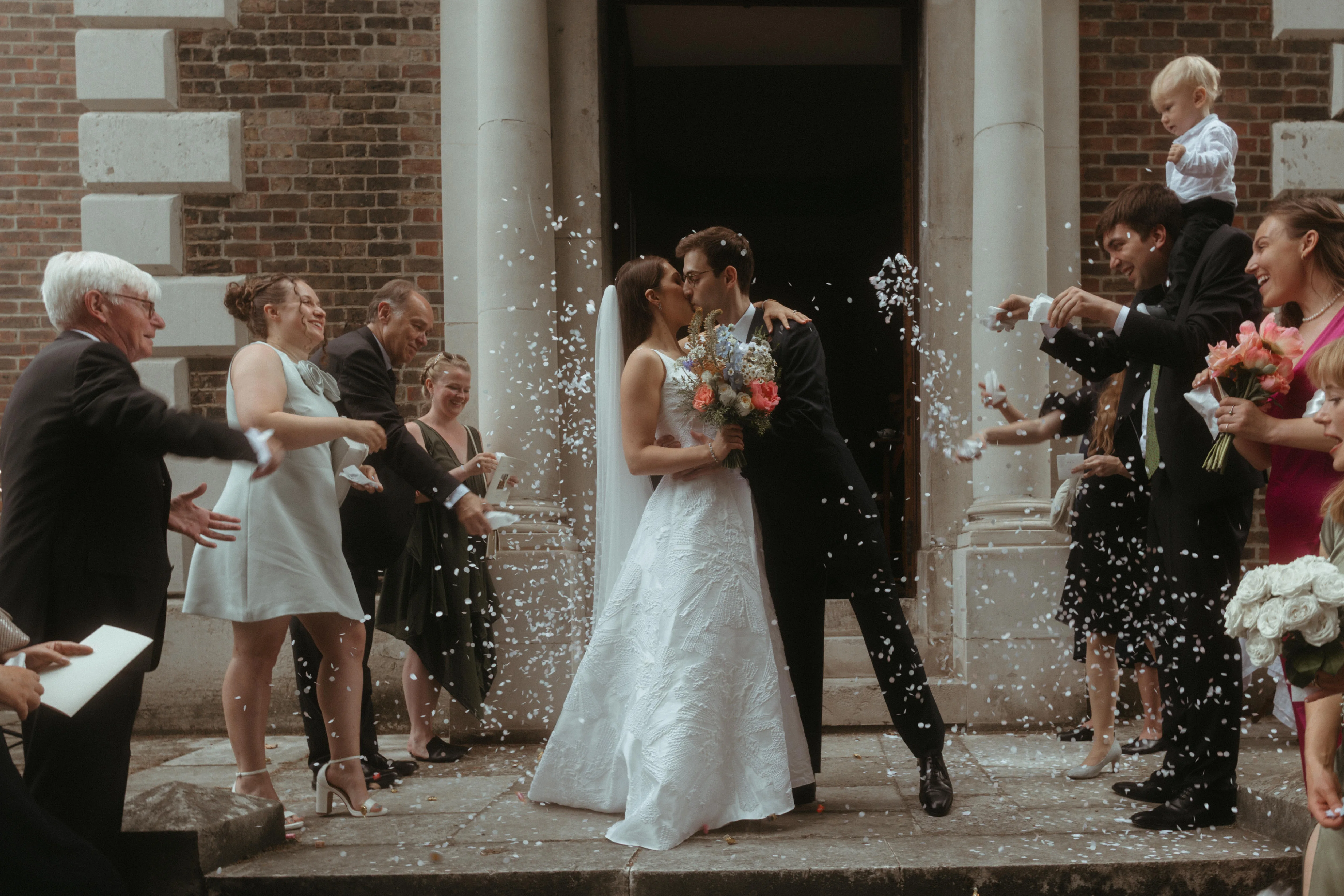 A bride & groom embrace outside a church as their friends and family shower them with confetti.