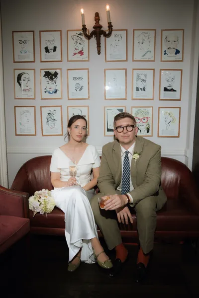 A bride & groom sit together on a leather sofa. They are enjoying a cocktail together after just getting married. The brides bouquet sits to her side.