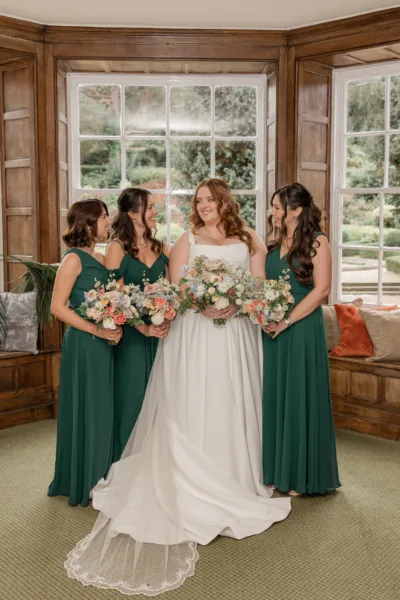 A bride stands with her three bridesmaids. All are holding their floral bouquets. The bride is dressed in ivory and the three bridesmaids are all wearing dark bottle green dresses.