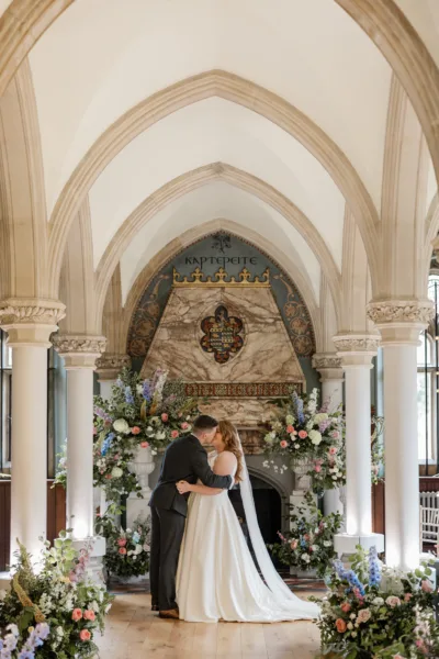 A couple embrace in The Library at Wotton House Hotel. Surrounded by overflowing garden urns and meadow style arrangements. Full of summer flowers.