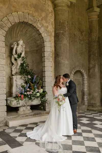 A bride & groom kiss whilst standing by an old stone fountain inside a folly at Wotton House Hotel. The old fountain behind is full of flowers, including roses and delphiniums. The bride is holding her bouquet.