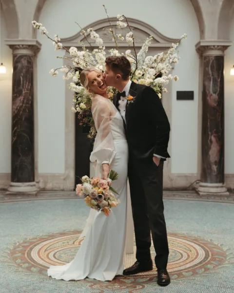A groom kisses his bride in front of a large urn full of spring blossom. The bride holds her bouquet.