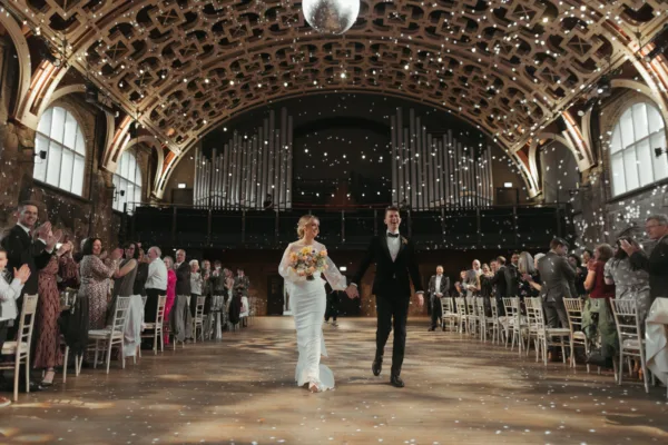A bride & groom walk through The Grand Hall at the Battersea Arts Centre. The Bride is holding her bouquet.