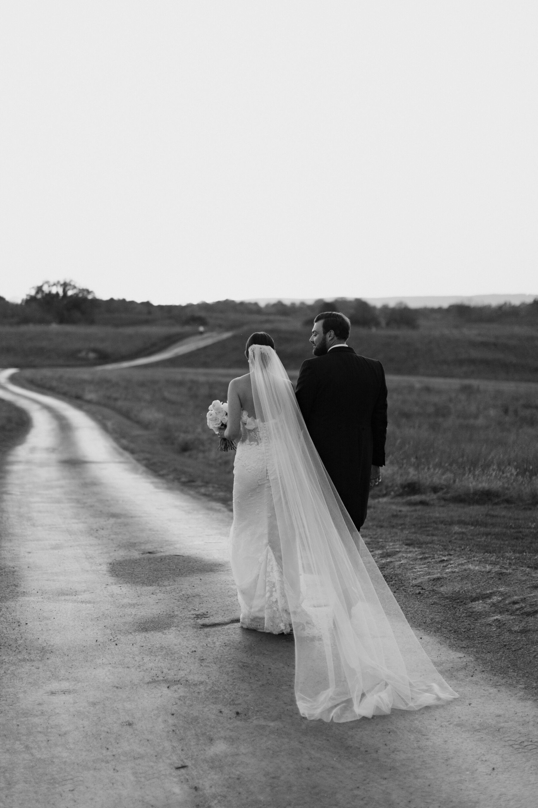 Shot behind of bride and groom walking along a path near fields. Brie wears a long wedding veil and carries a white bouquet. Groom wears traditional tails.