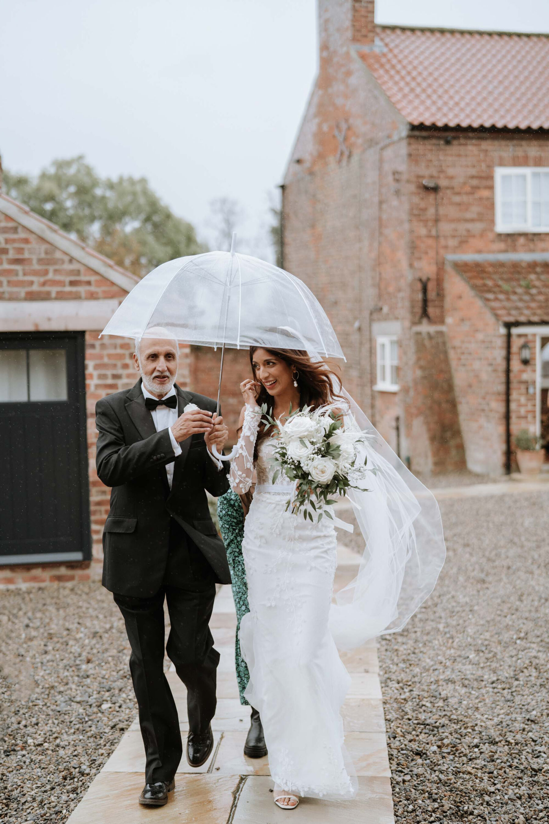 Bride arriving with the father of the bride in black tie and holding an umbrella.