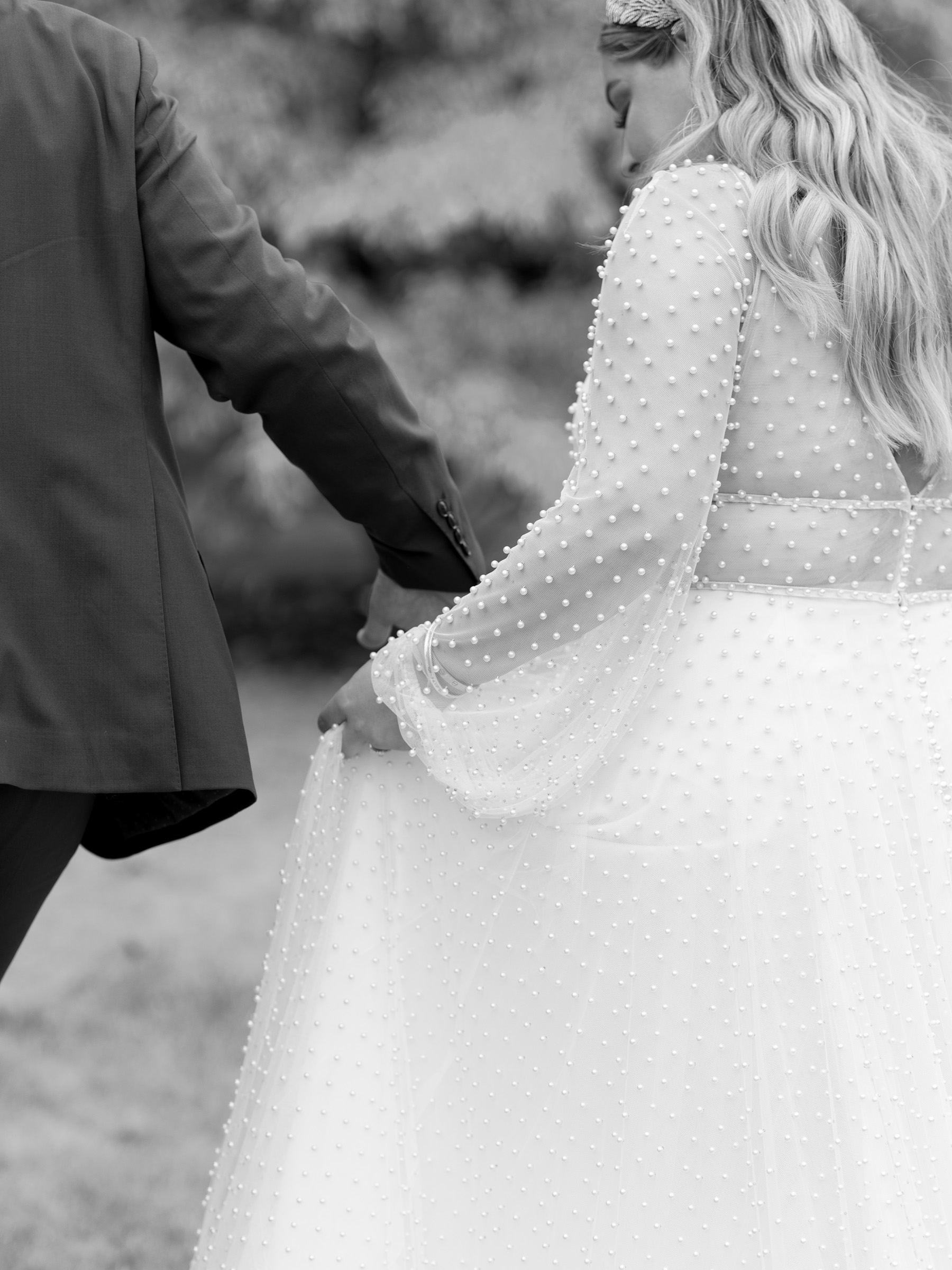 Black and white photograph of bride in a pearl wedding dress holding hands with her groom.
