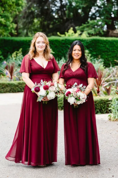 Two bridesmaids wearing deep burgundy dresses holding their bouquets whilst stood outside Pembroke Lodge, Richmond Park.