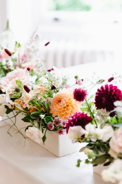 A low ceramic vase sits on a table full of dahlias in orange and burgundy, with other seasonal flowers.