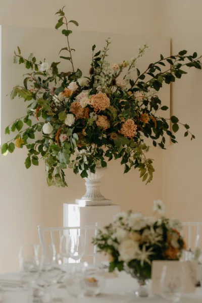 A large urn full of seasonal foliages with toffee colours chrysanthemum and roses sits in a corner of a room.