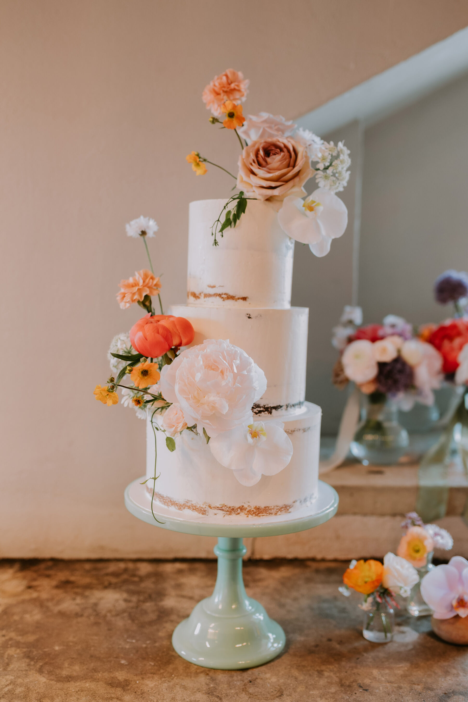 3 tier wedding cake on a pale green stand with colourful wedding flowers.