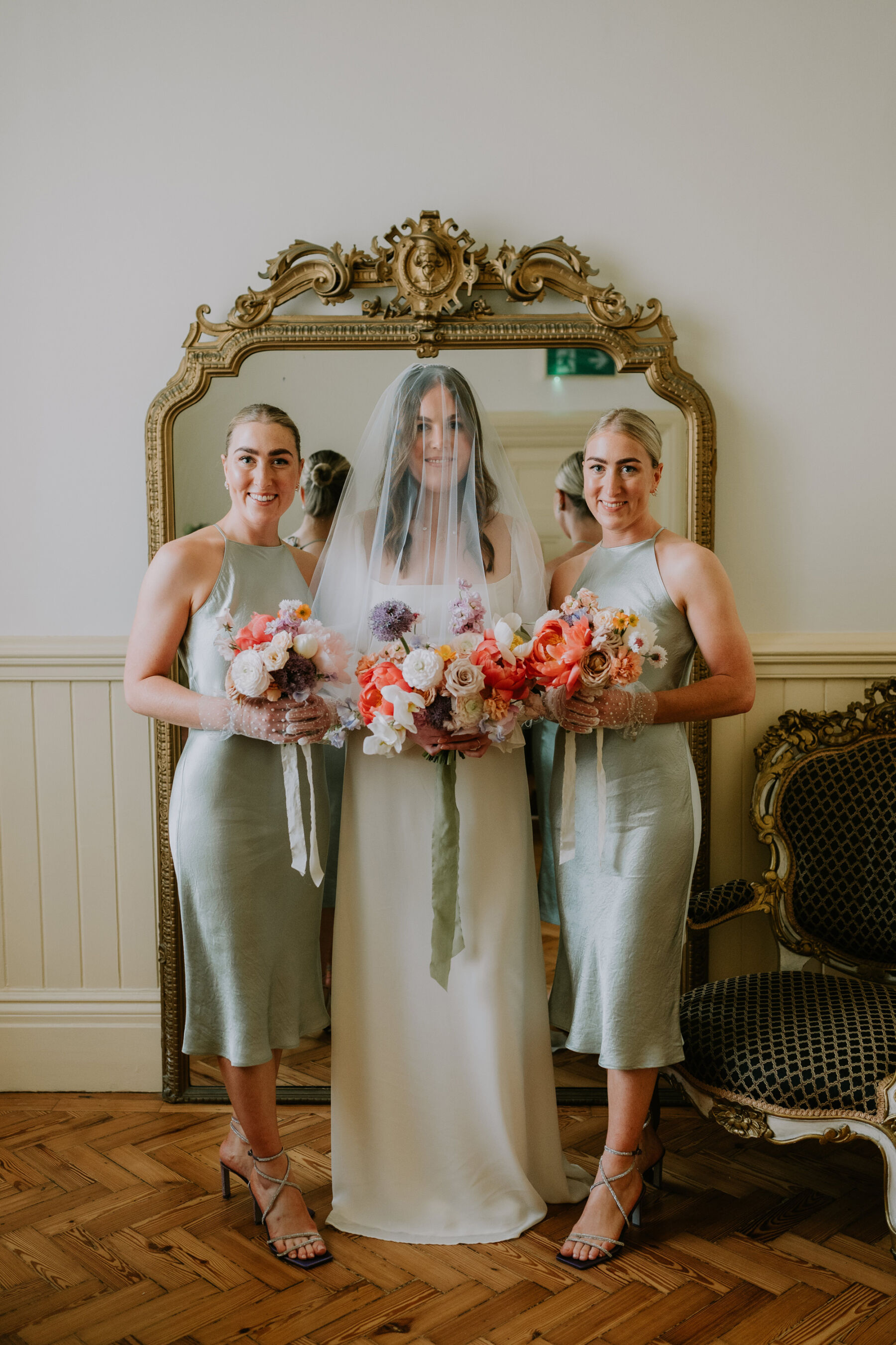 Bridesmaids in calf length pale green halterneck slip dresses and carrying colourful bouquets. Bride wears a Reformation dress and single tier veil over her face.