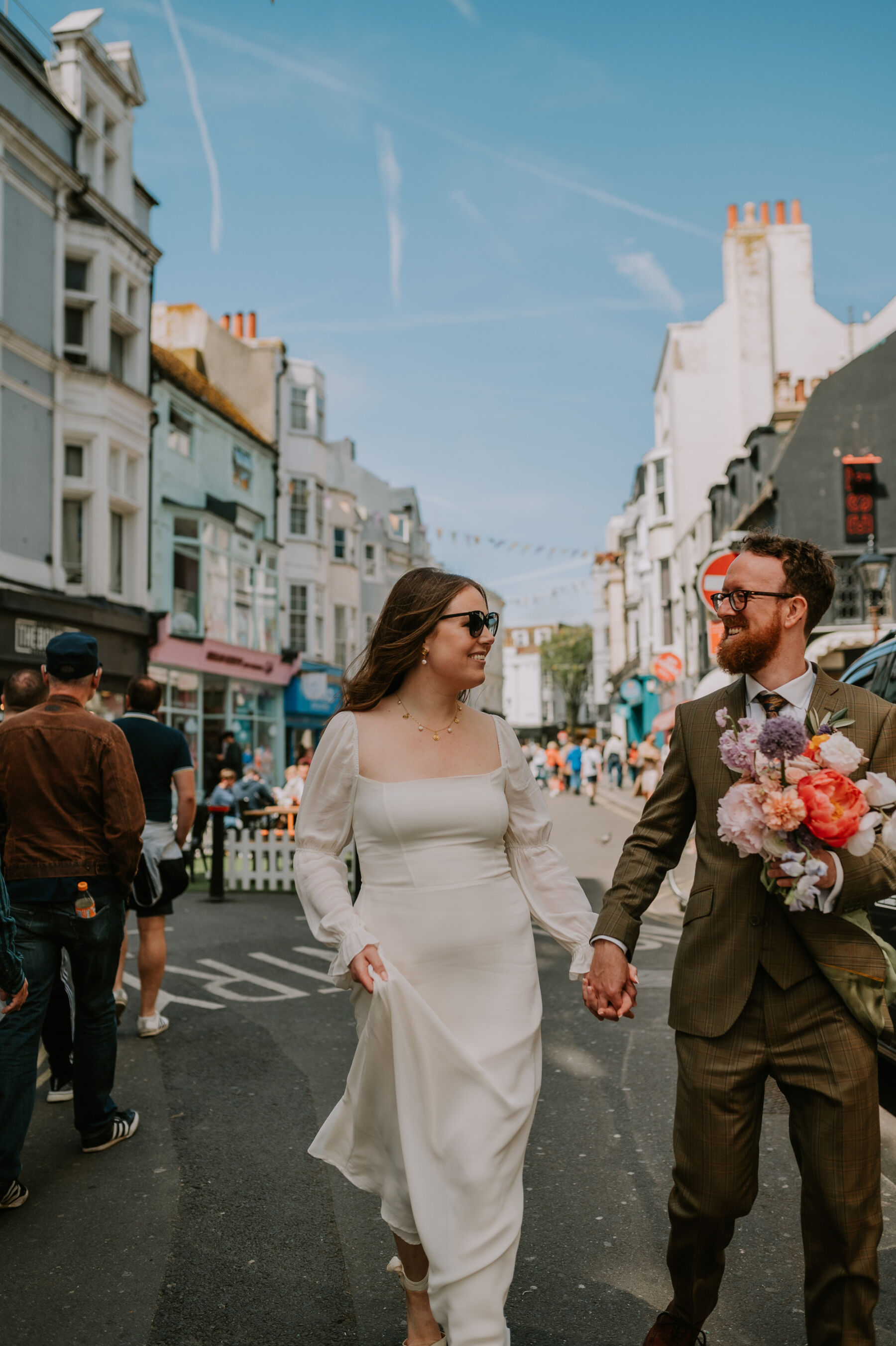 Bride and groom walking through Brighton. Bride wears a Reformation dress. Groom carrying the bride's colourful bouquet.