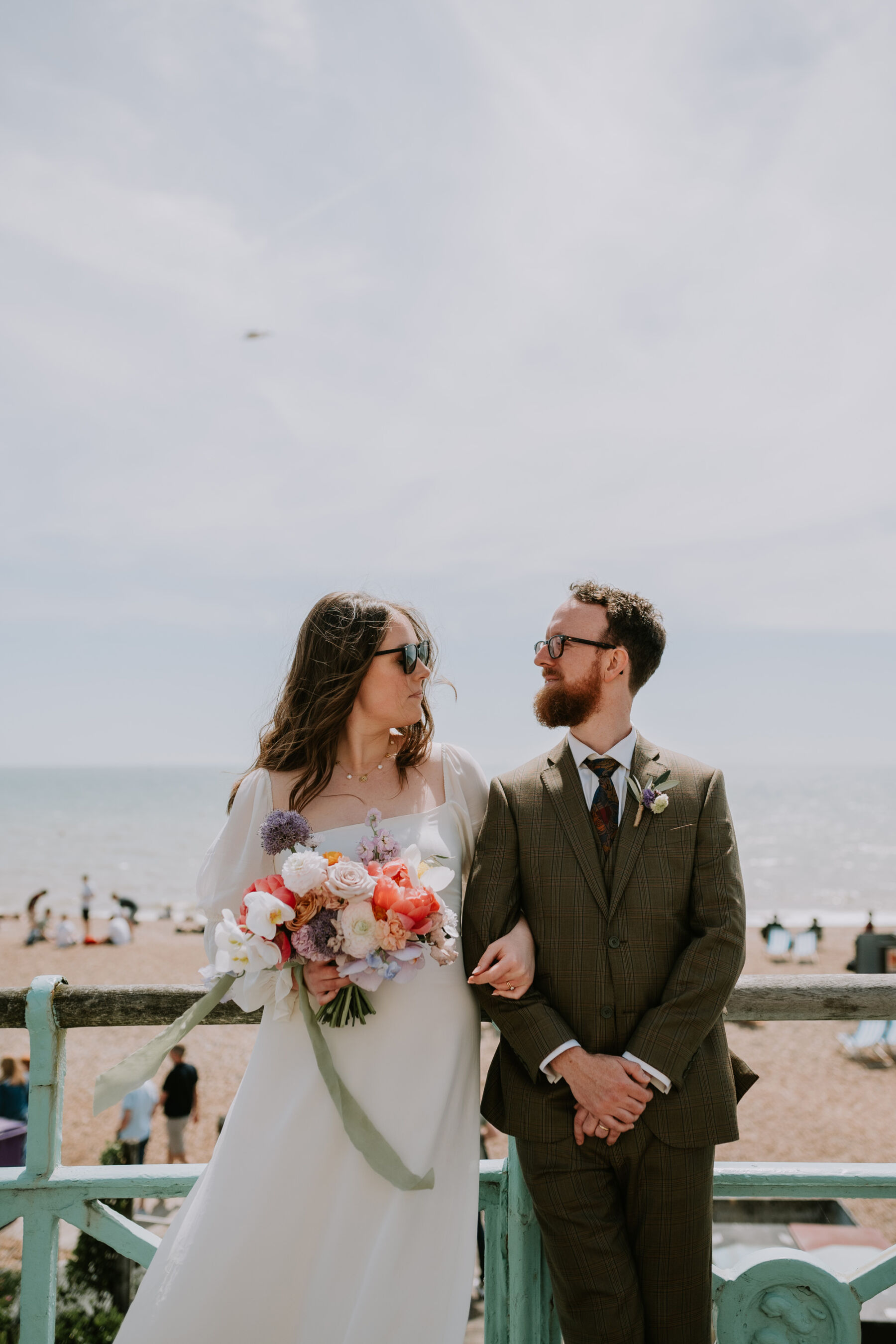 Bride and groom standing by the seaside in Brighton. Reformation wedding dress. Colourful bouquet with pale green ribbon.