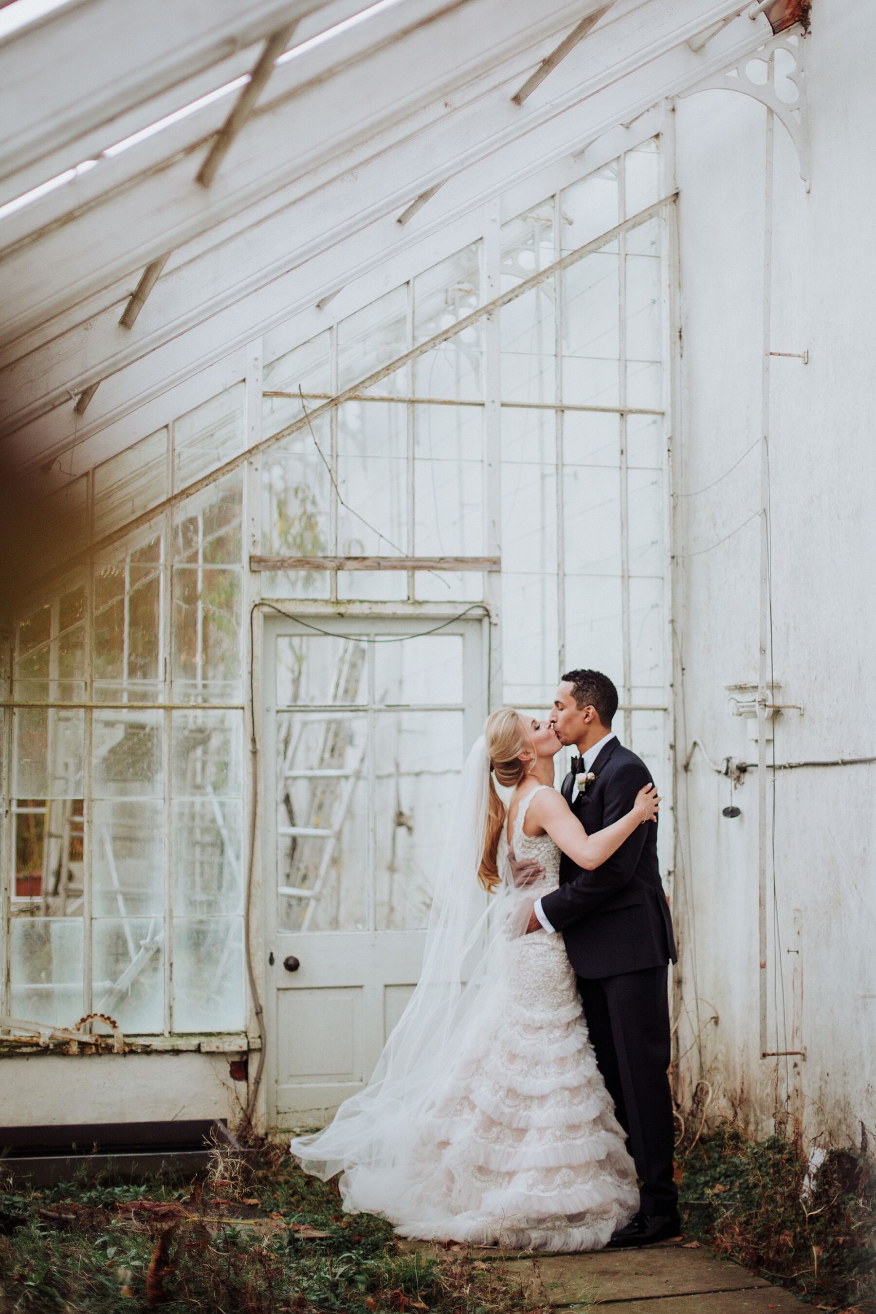 Bride standing by a glasshouse wearing a tiered wedding dress, by Claudia Rose Carter Photography.