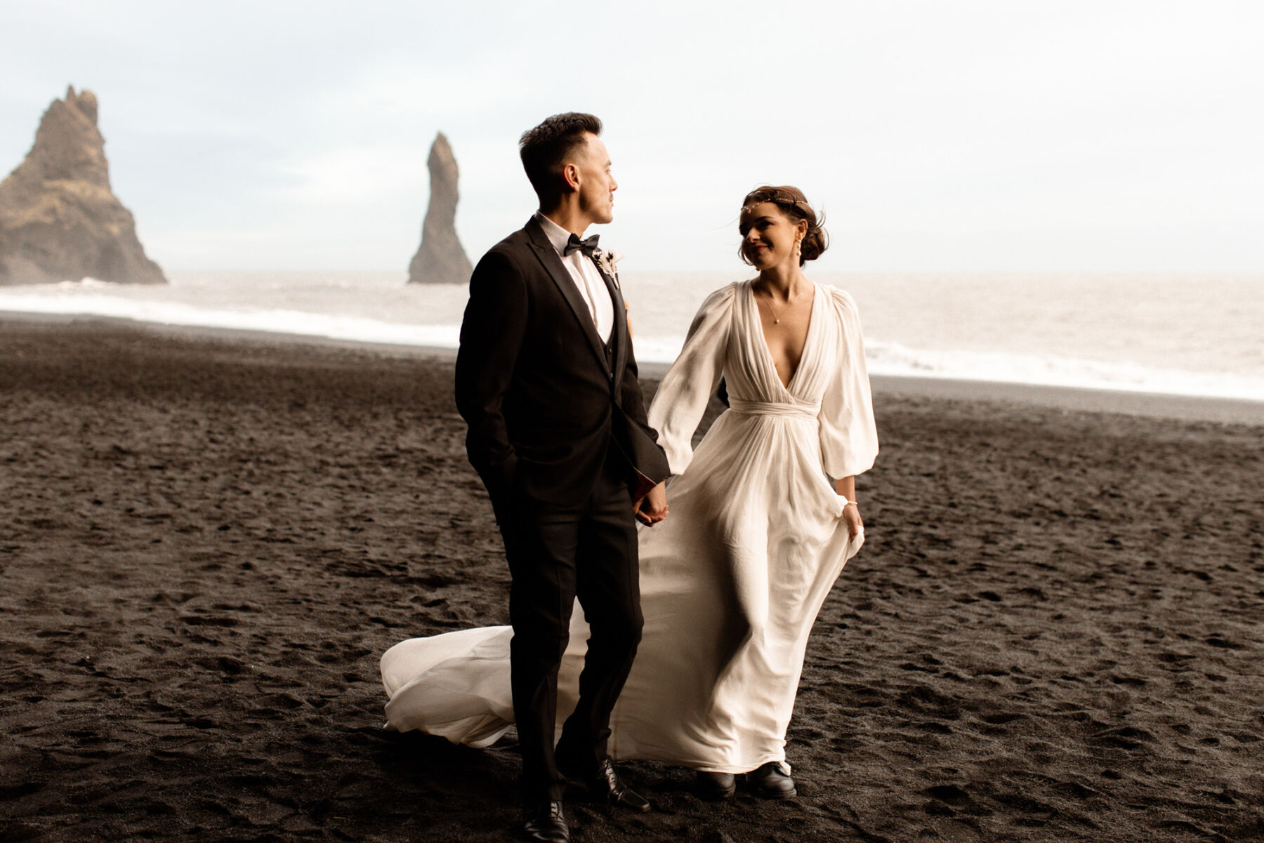 Windswept scene on a black sands beach in Iceland. Groom in black tie, bride in billowing silk dress