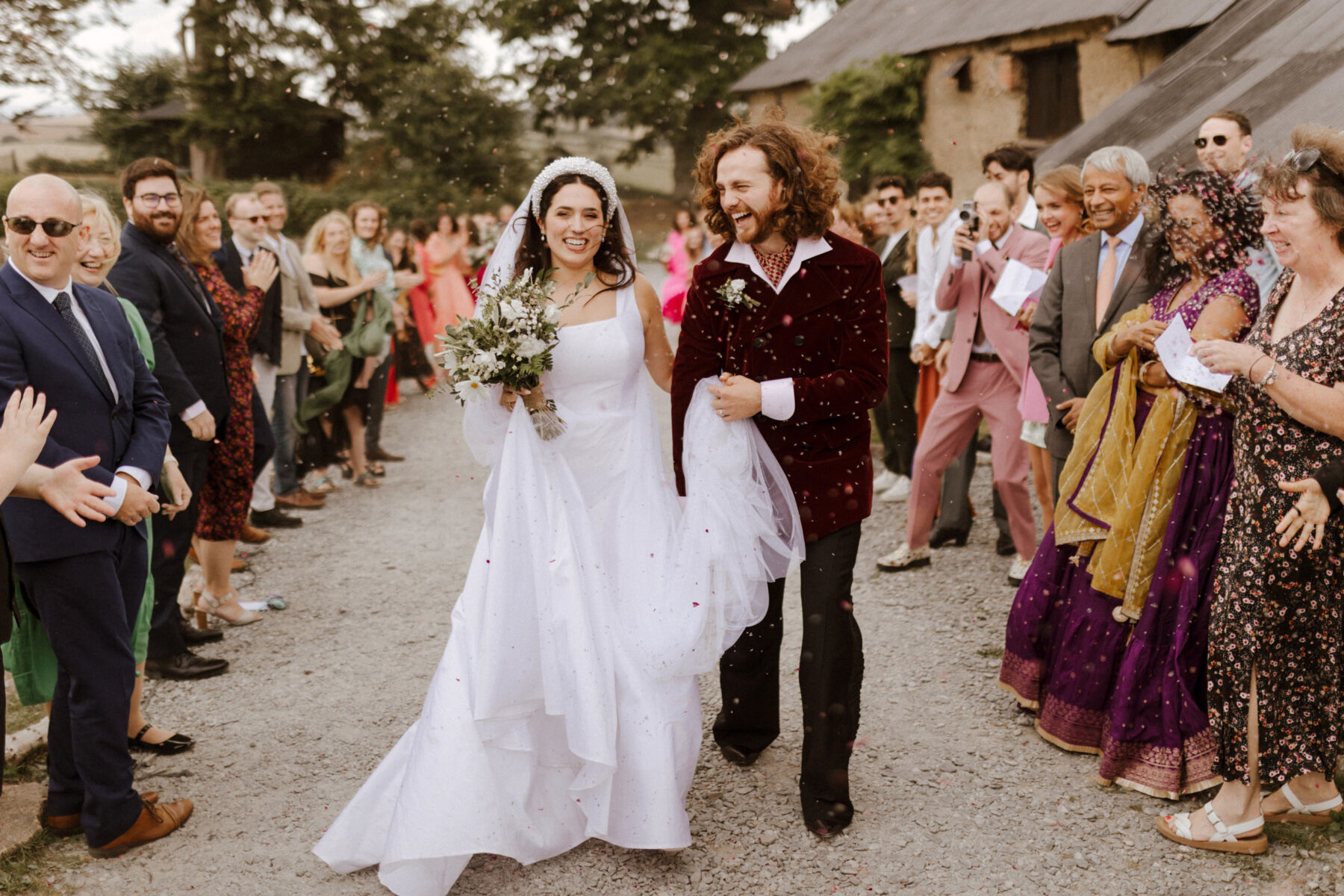 Very happy bride and groom in a shower of confetti. Bride hooks her arm around the groom, who is wearing 70's inspired flares and a dark red velvet jacket. Bride wears Jessica Bennett dress, pearl headband, veil and carries a bouquet of all white flowers.