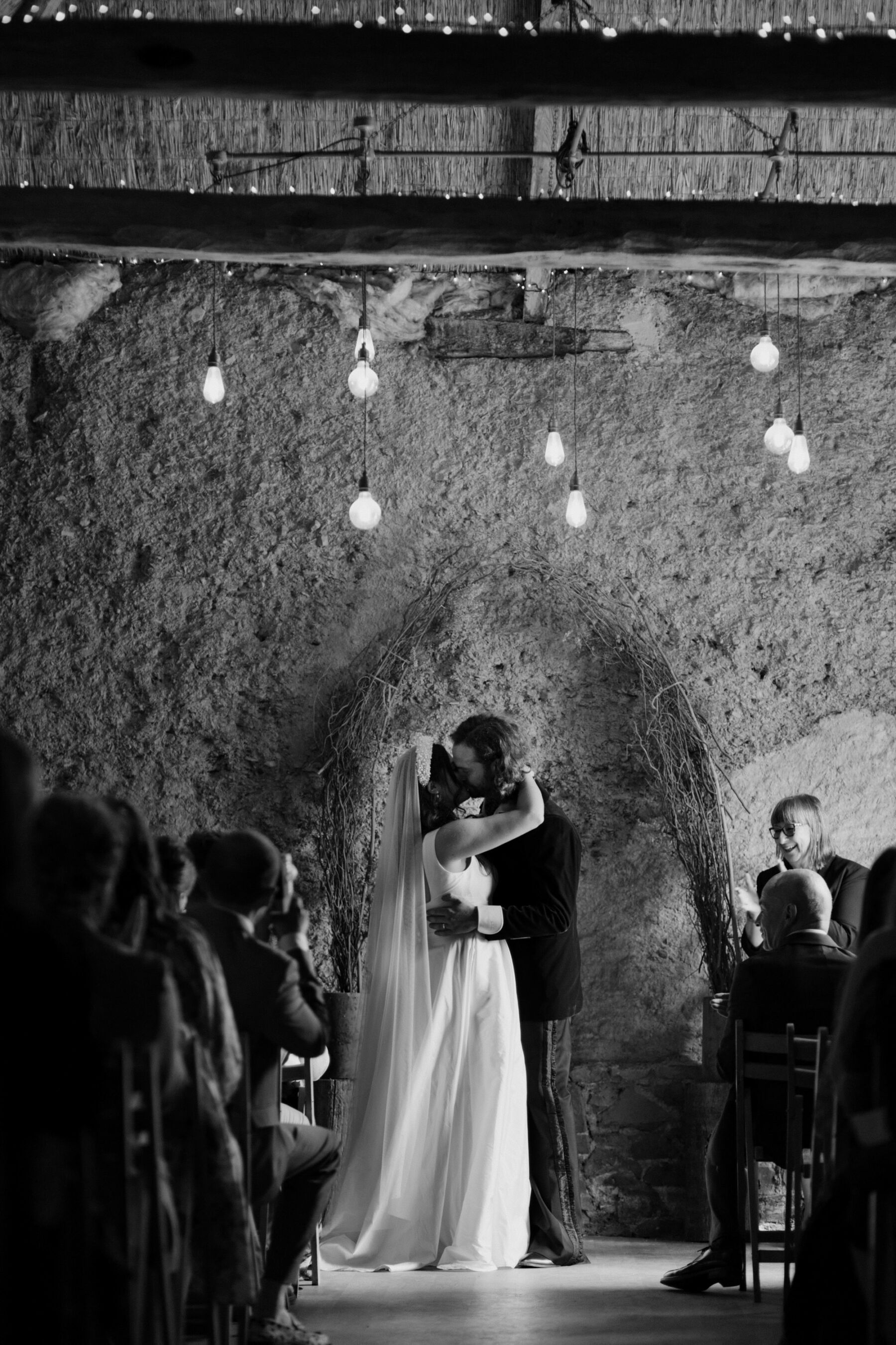 Bride and groom kissing at the end of their wedding ceremony whilst standing beneath festoon lights.