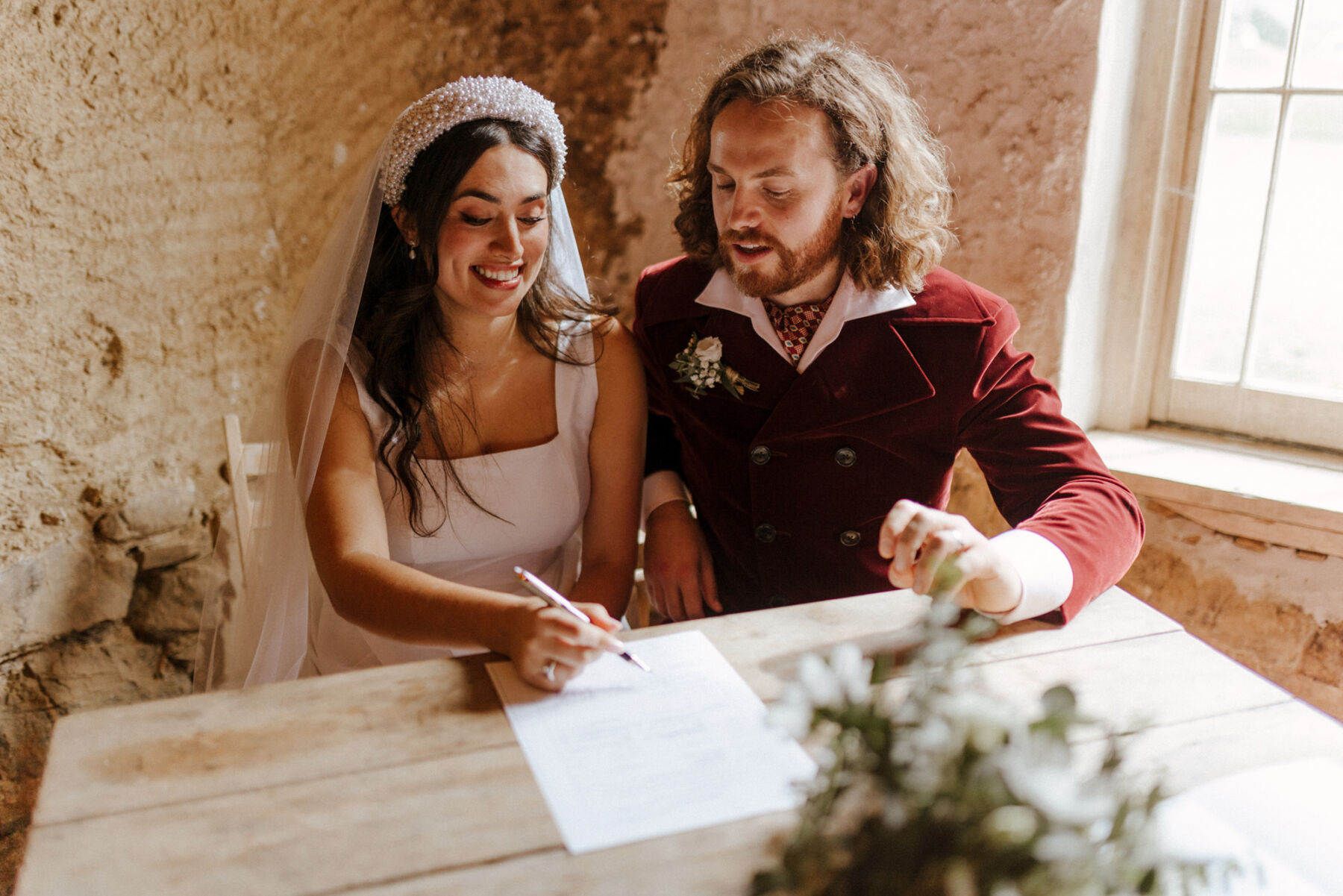 Cool bride and groom seated smiling and signing the register during their wedding ceremony.
