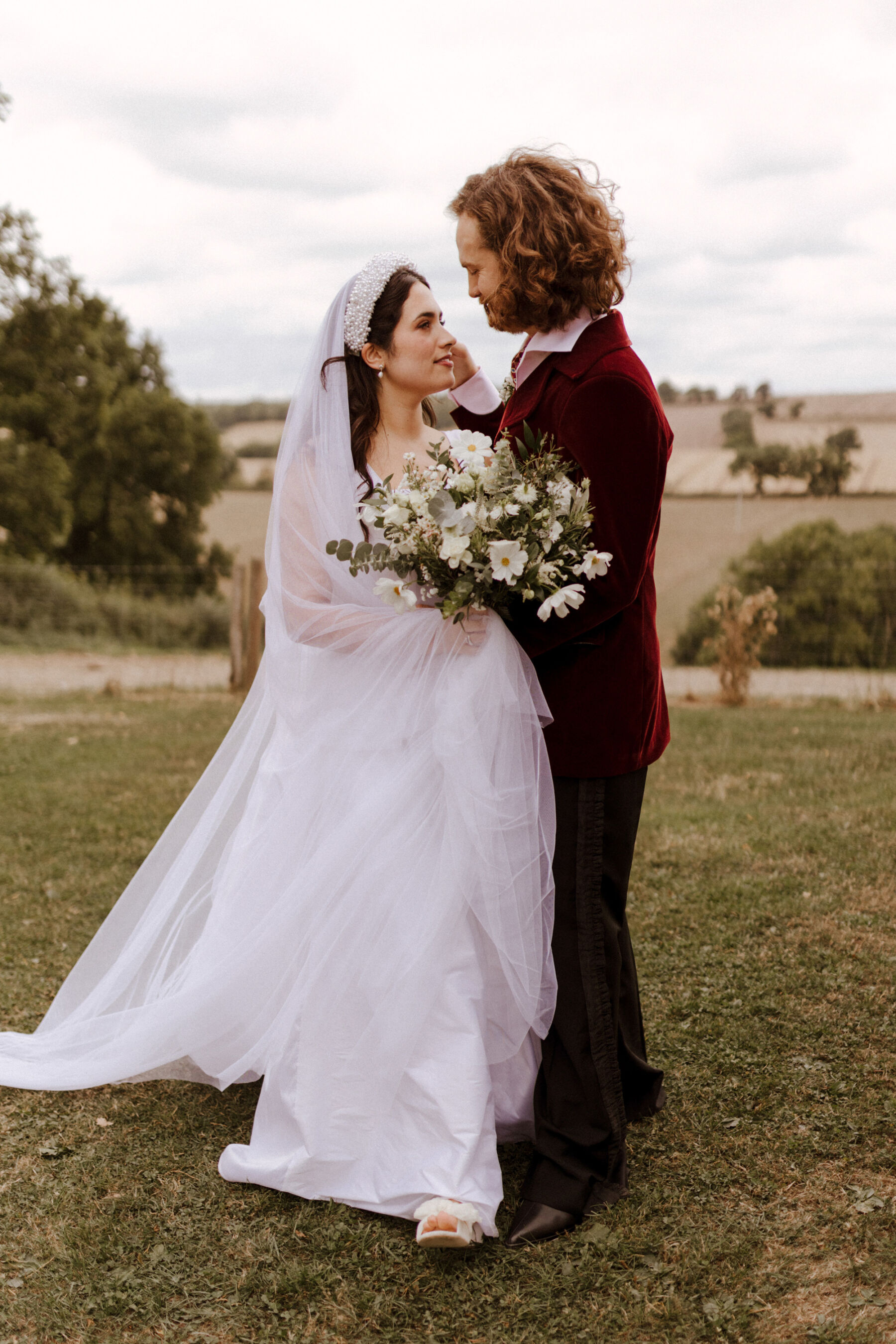 Groom in a maroon velvet suit lovingly touching the side of his bride's face. Bride wears a pearl headband , holds a bouquet of all white flowers and wears a Jessica Bennett wedding dress. 