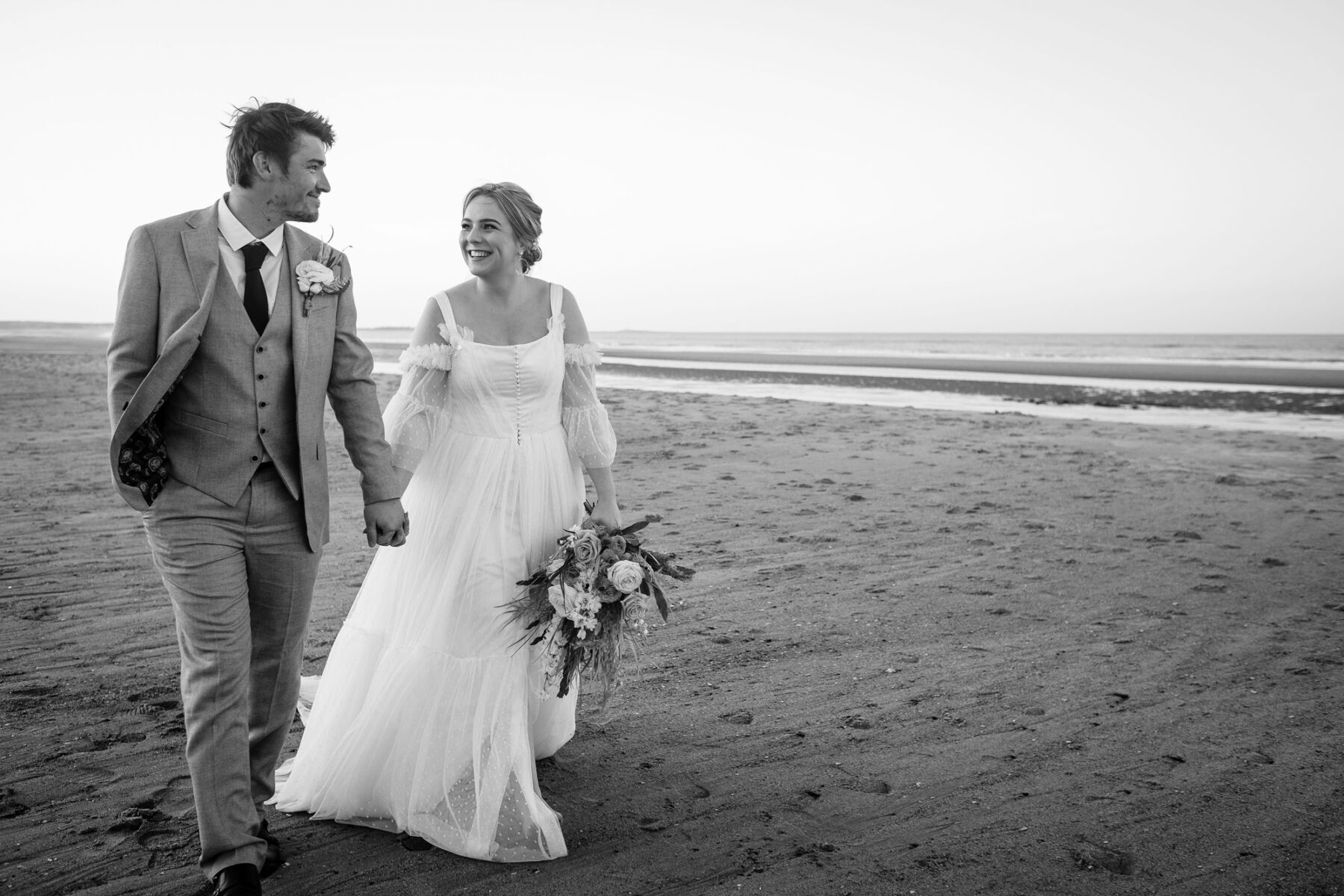 Bride and groom walking on a beach, bride wears a Kate Clare Couture wedding dress and holds a bouquet, looking lovingly into the eyes of her groom