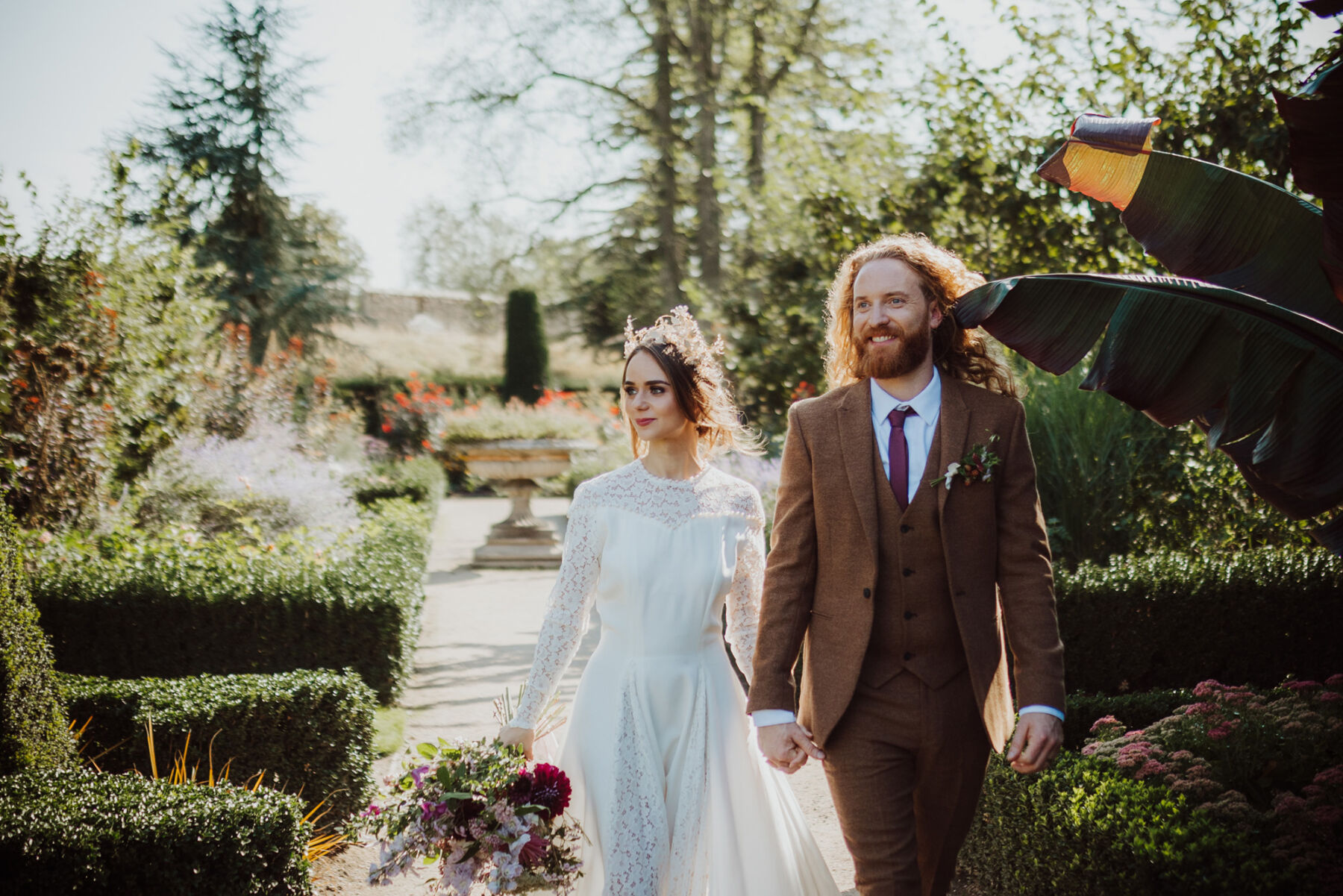 Bride wearing a vintage lace wedding dress and a bridal crown. Groom in a brown suit. Gardens at Bishop's Palace historic wedding venue in Wells, Somerset.