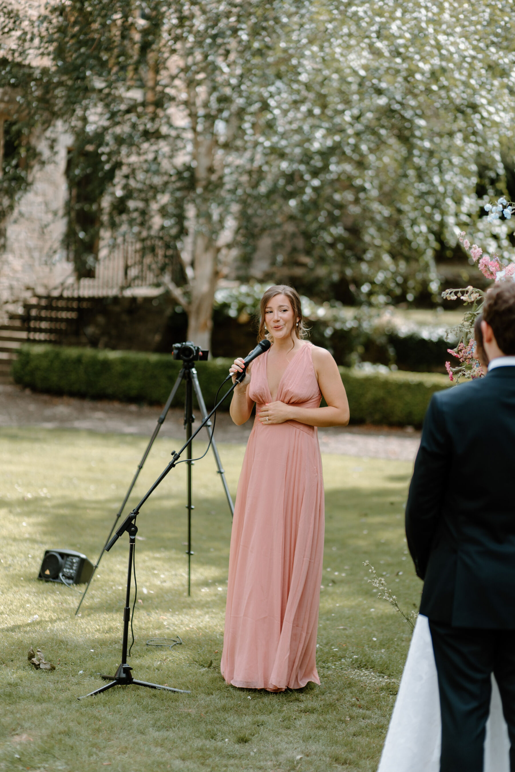 Bridesmaid in pink dress at Durham Castle speaking into a microphone during an outdoor wedding ceremony.
