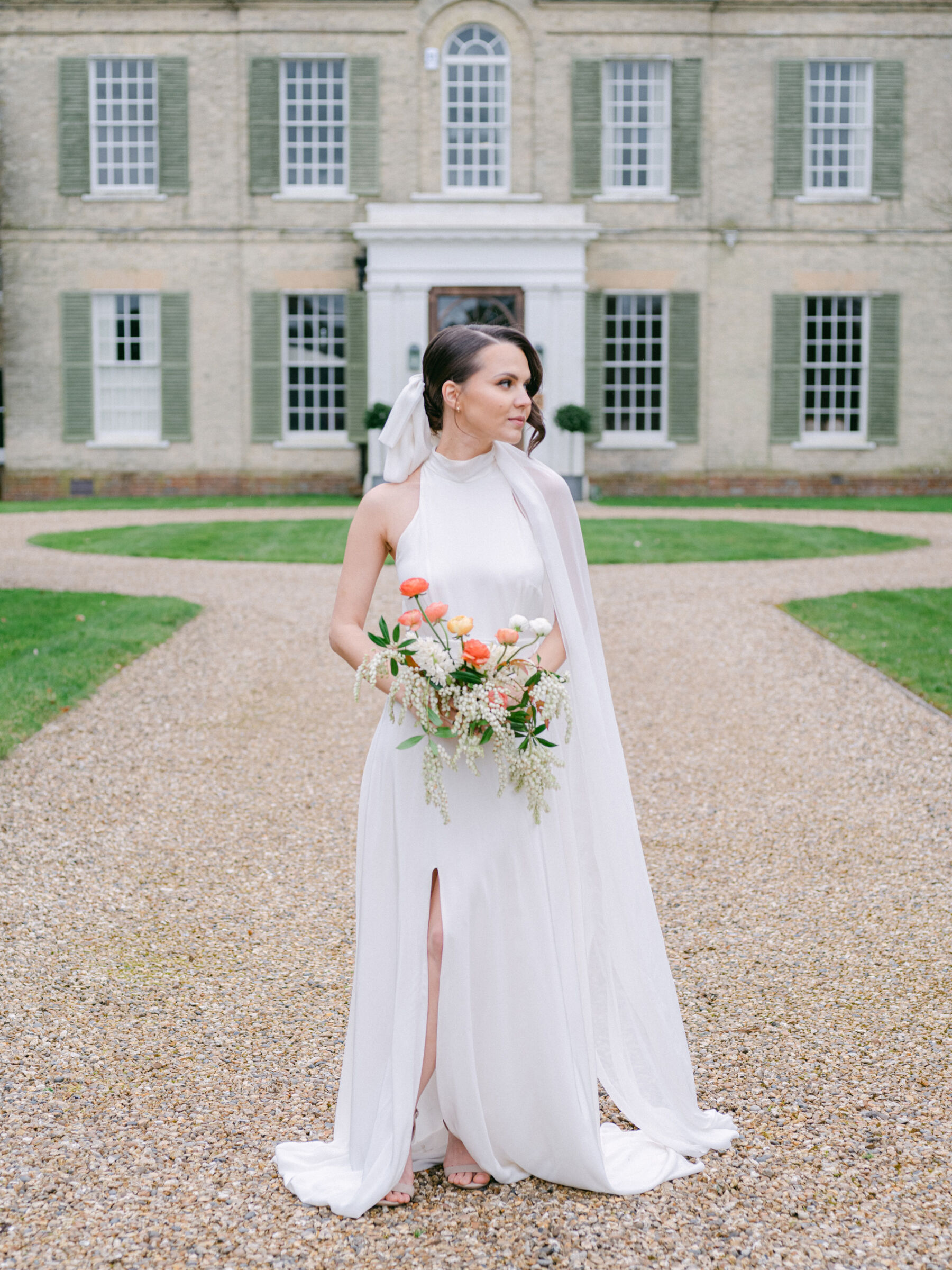 Bride standing in the grounds of Findon Place wedding venue wearing a halterneck wedding dress with a thigh length slit. The bride carries a bouquet of British grown Ranunculus and seasonal flowers.