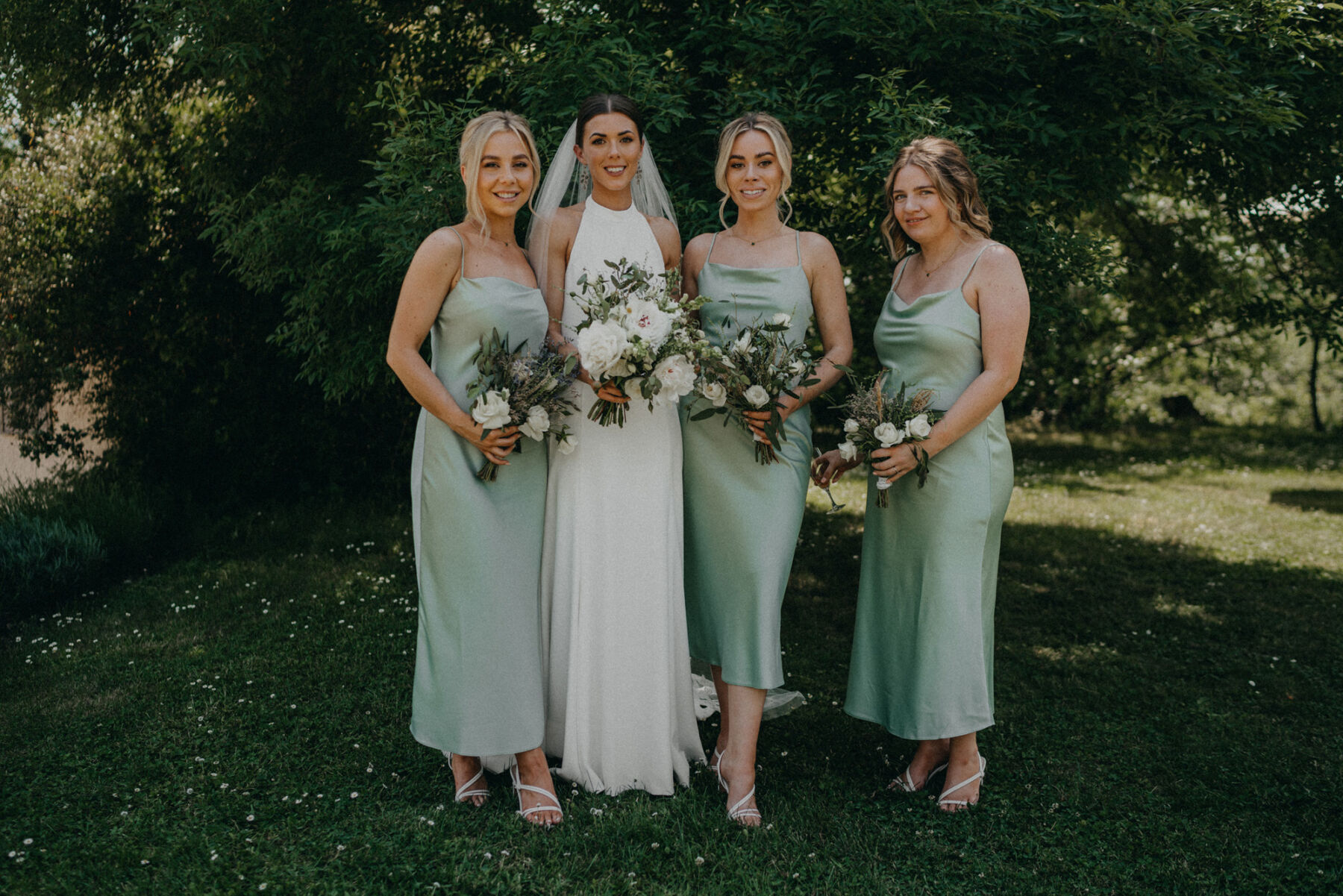 Bride in a halterneck wedding dress with her bridesmaids in pale green dresses holding all-white bouquets.