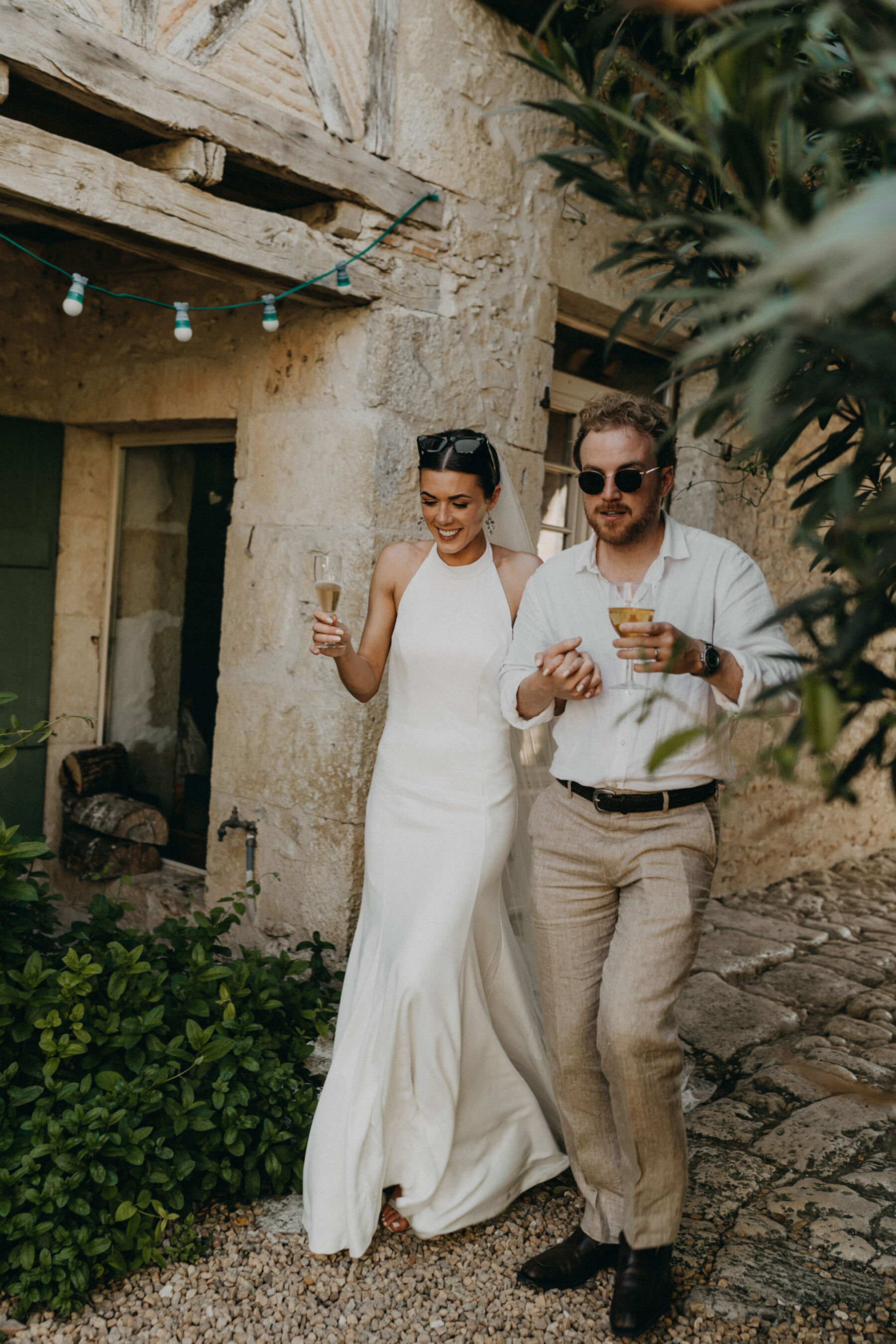 Bride in a halterneck wedding dress holding a glass of chapaign with her groom in a linen shirt and beige trousers.