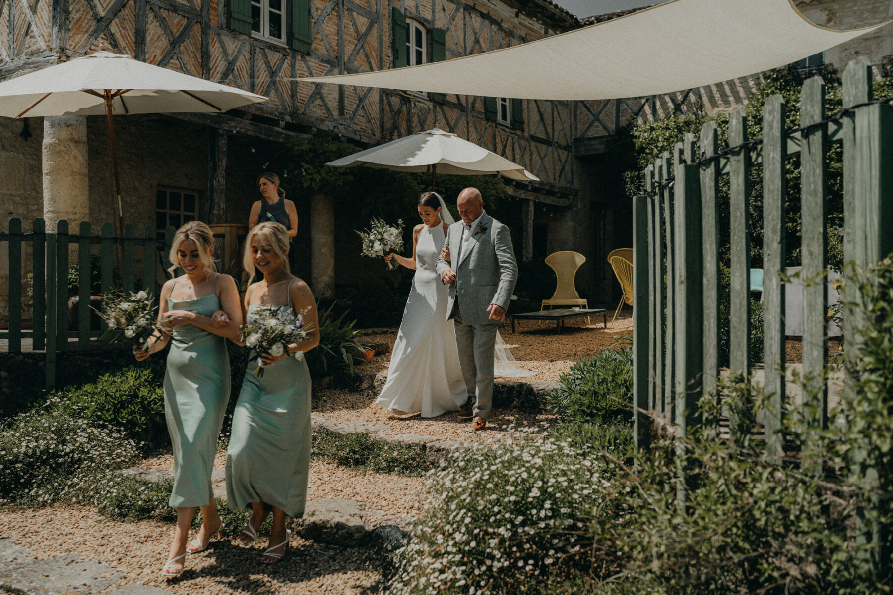 Emily in a Bespoke Halterneck Gown for her Elegant Chateau De Puissantut Wedding in South West France 14 Bride in a halterneck wedding dress in arms wtih her father making her way to the wedding ceremony with her bridesmaids in pale green dresses.