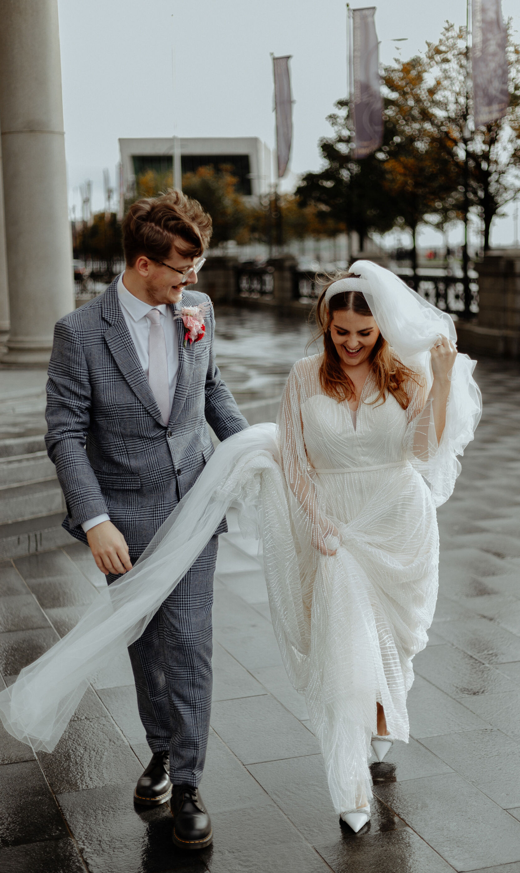Bride & Groom in Liverpool city. Bride wears a sequin wedding dress, pearl headband, pointed toe shoe and long veil that is being gathered by the groom.