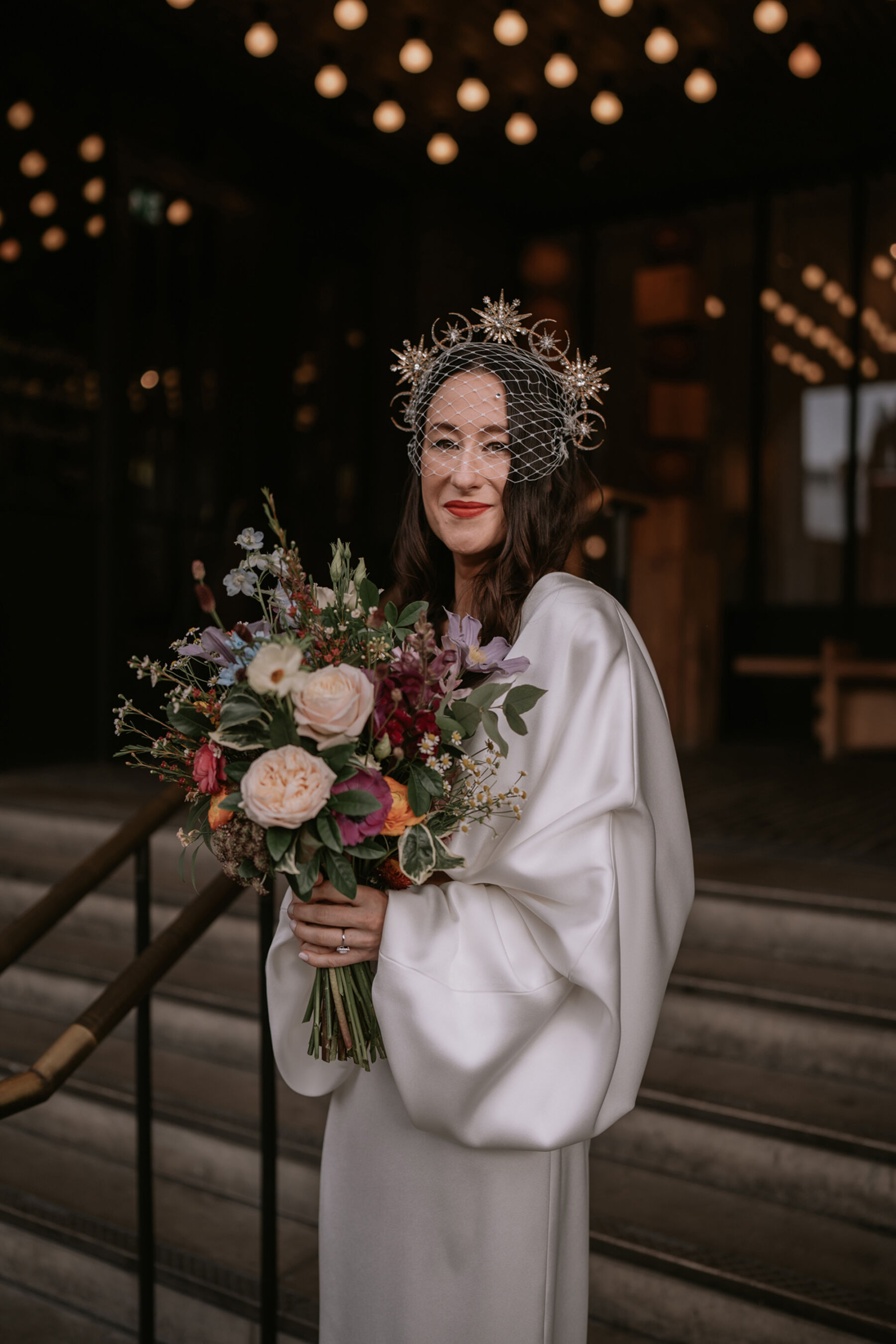 Bride in a modern and ultrastylish Bon Bide wedding dress with long billowed sleeves, carryinga. colourful modern bouquet and wearing a birdcage veil.