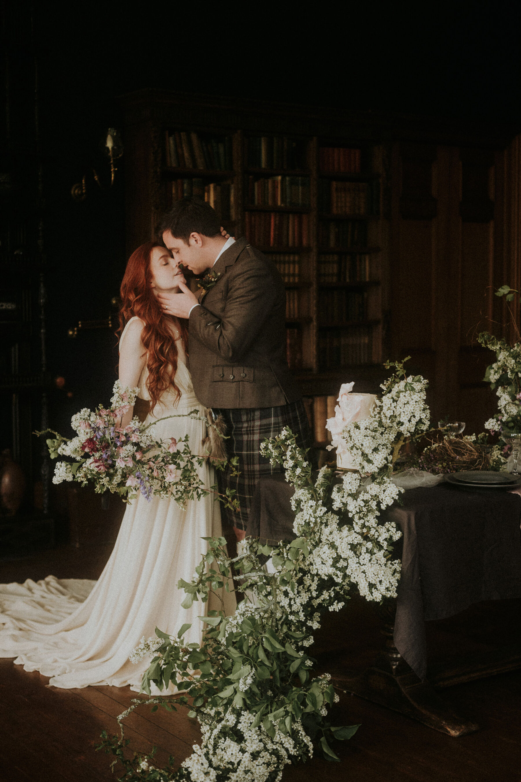 Bride with long red hair being held and kissed by her groom. Beautiful neutral white flowers surround them.