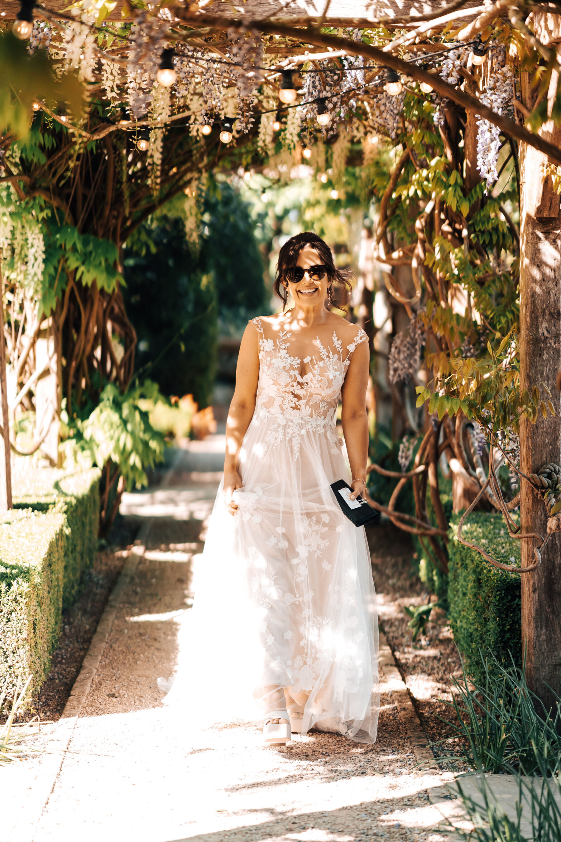 Bride in an Anna Kara dress and sunglasses walking beneath the wisteria, at Tythe Barn wedding venue in the Cotswolds.