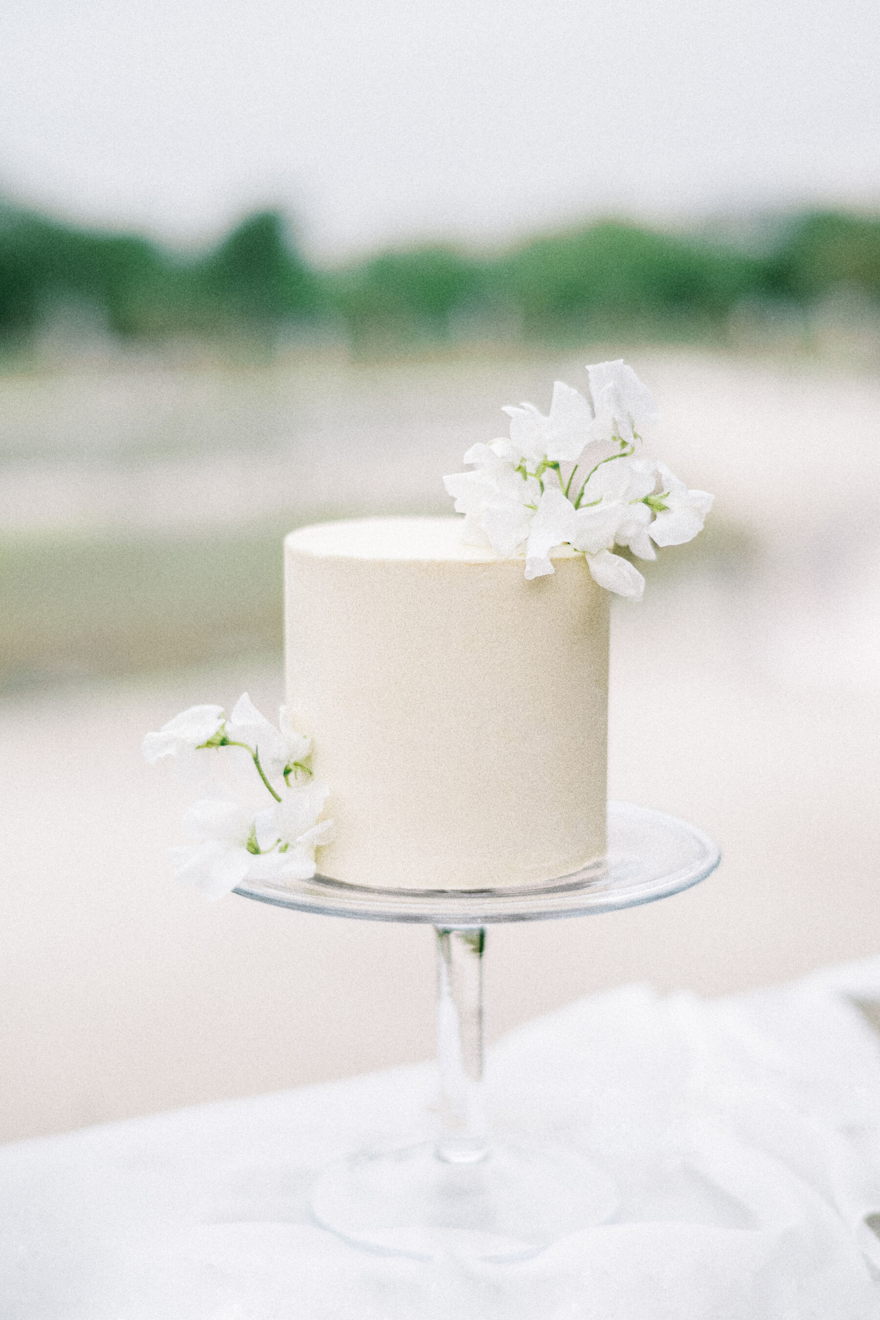 Single tier wedding cake with white sweet peas on a tall glass cake stand.