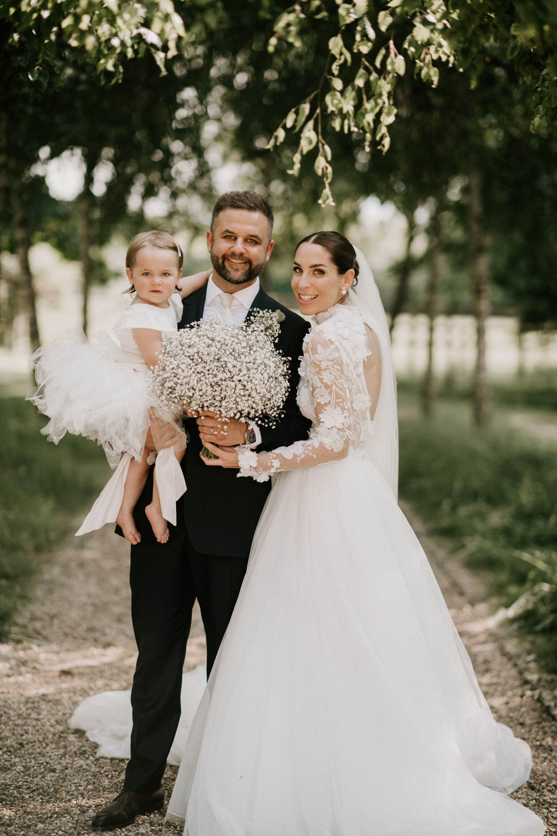 Bride wearing a J Andreatta dress standing with her groom who is holding a flowergirl in his arms.