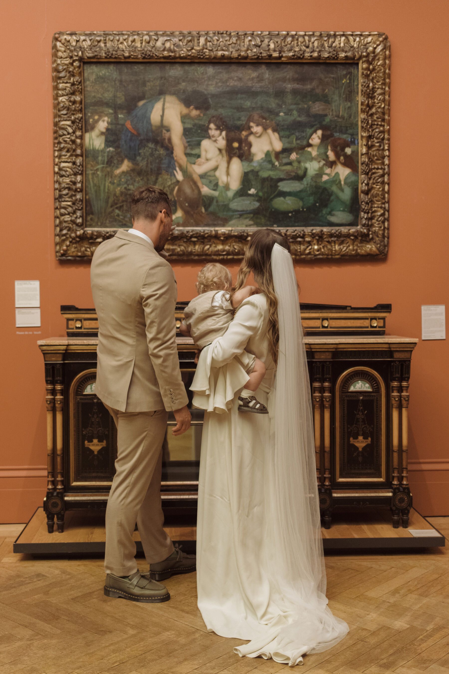 Bride and groom in Manchester Art gallery looking at a painting. The bride is holding her baby boy in her arms and wears a Ghost wedding dress. The groom wears a pale suit.