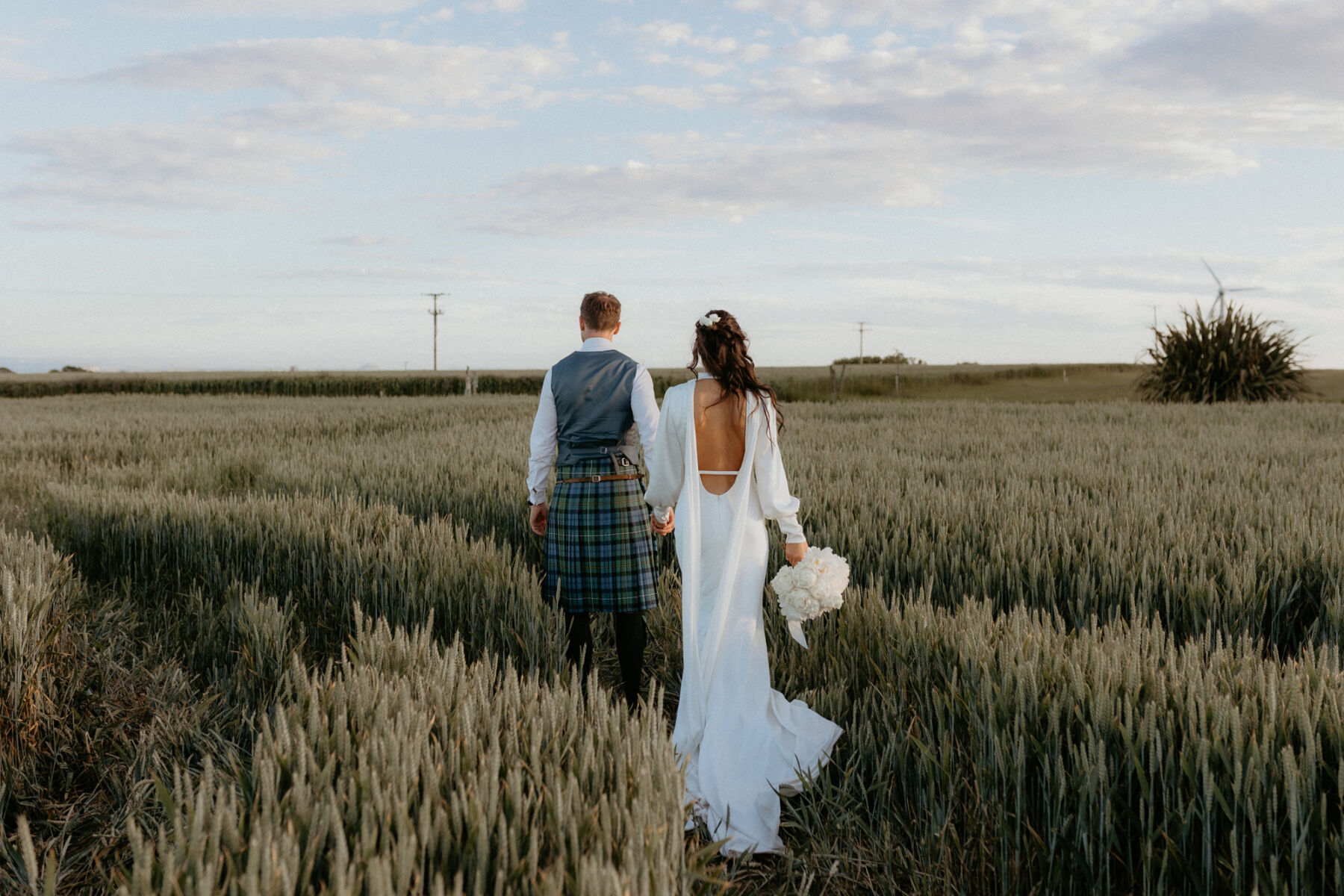 Bride in a backless wedding dress walking with her groom through a field