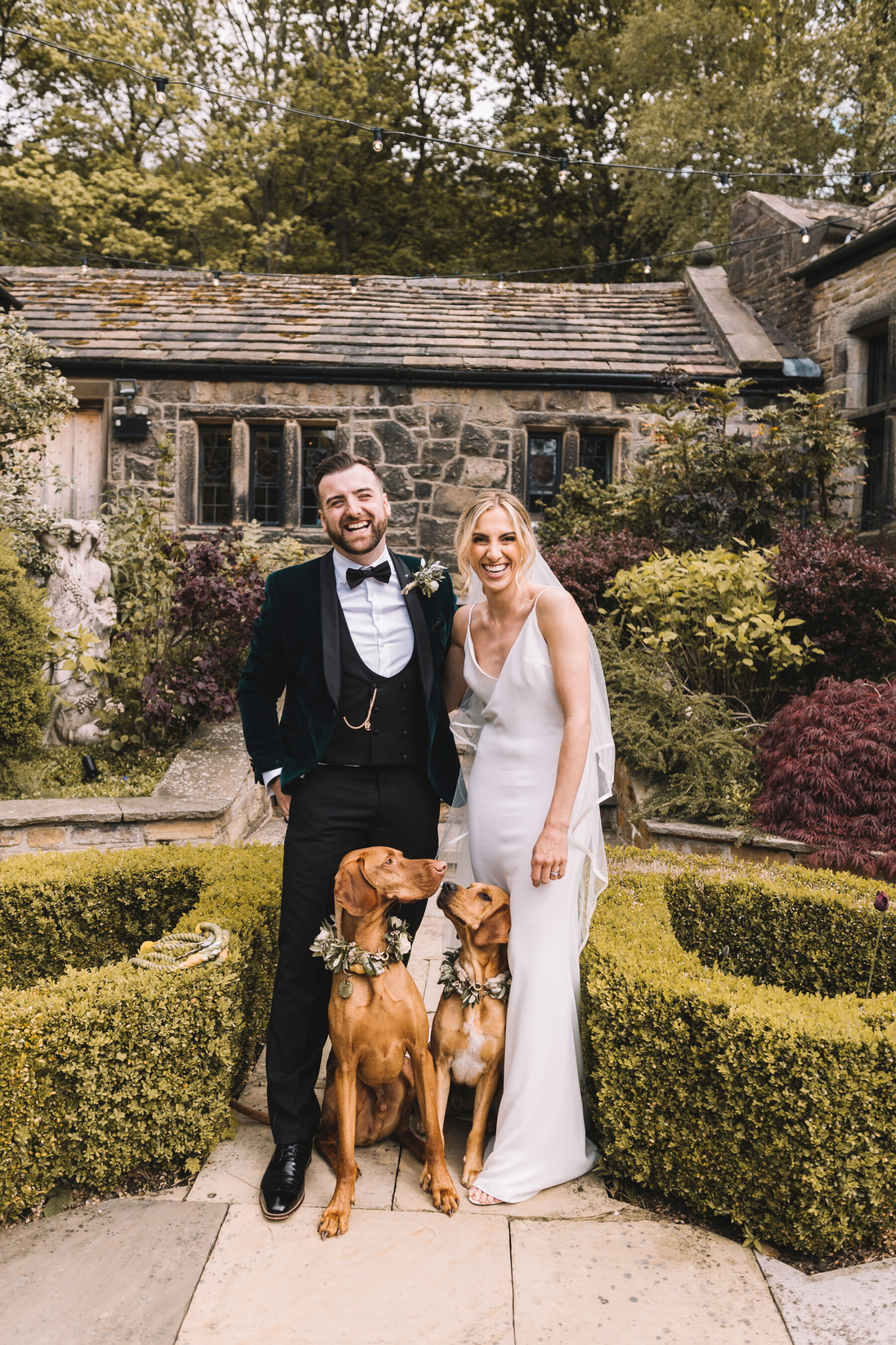 Bride and groom in black tie at Upper House in Hayfield, standing with their 2 Vizsla dogs wearing floral collars.