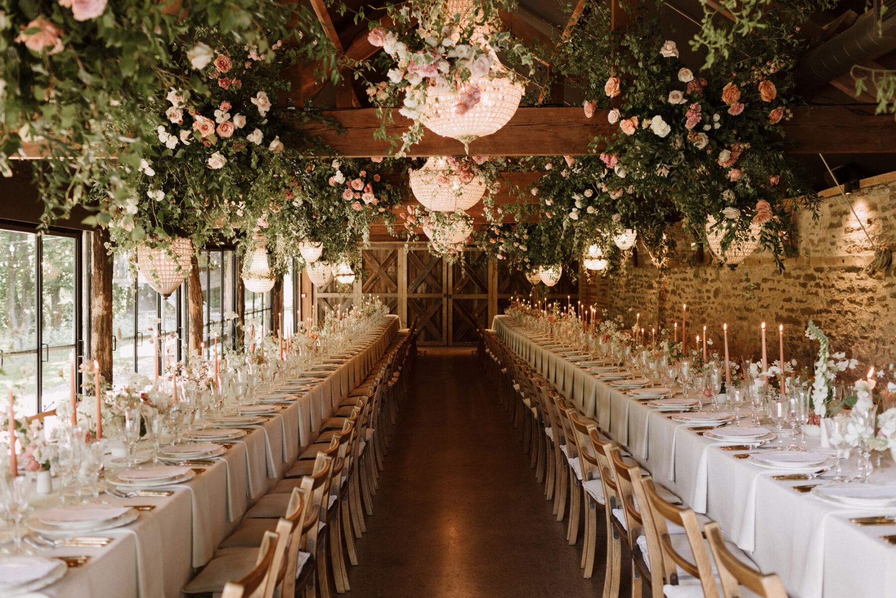 Inside the barn at Dewsall Court, country house wedding venue in Herefordshire. Trestle tables and taper candles with hanging greenery and chandeliers hanging above the tables.